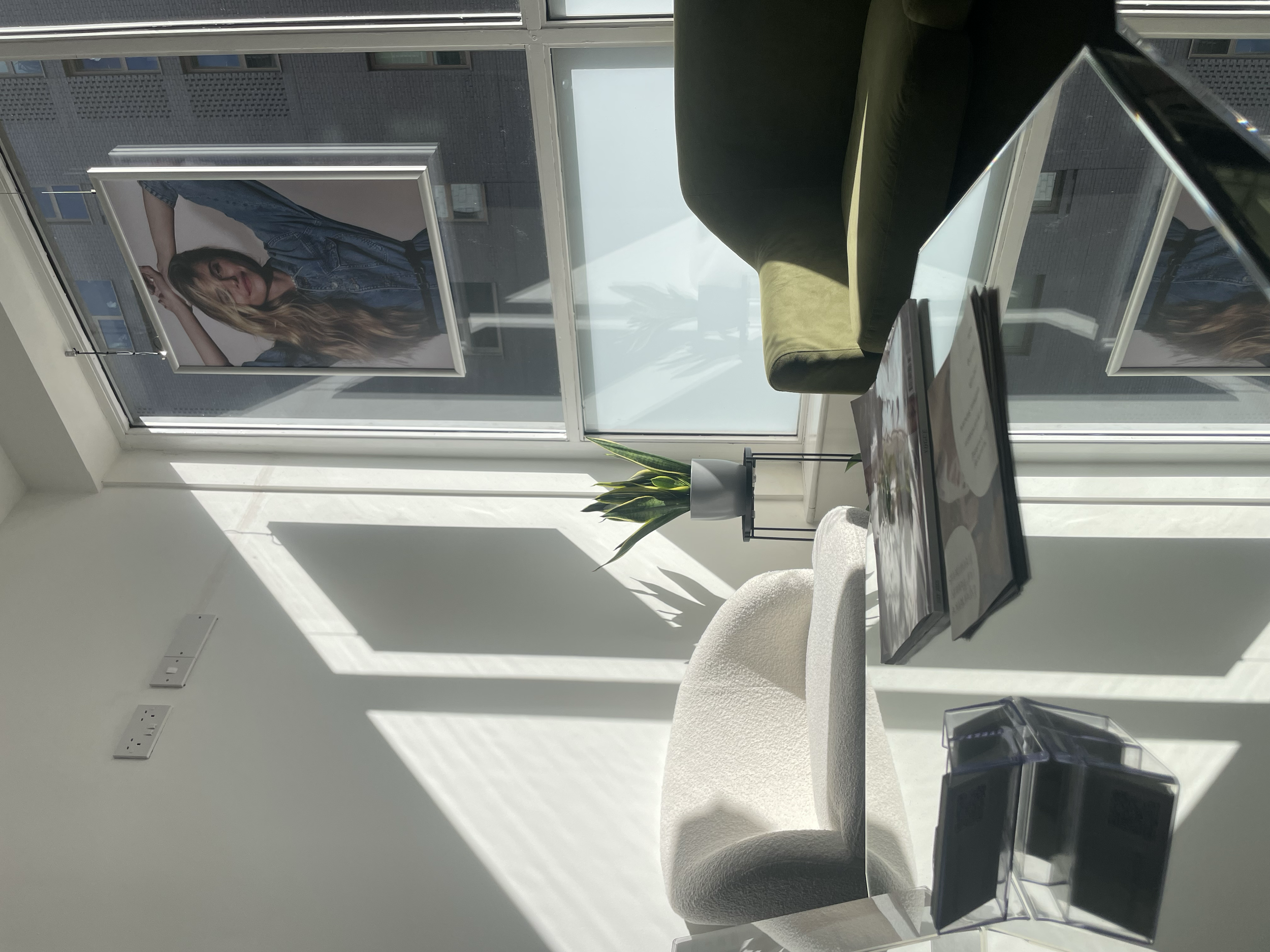 Bright office waiting area with a large window, a green sofa, a plant on a stand, a white armchair, a laptop on a table, and framed photograph of a woman on the window. Shadows from blinds are cast on the wall.