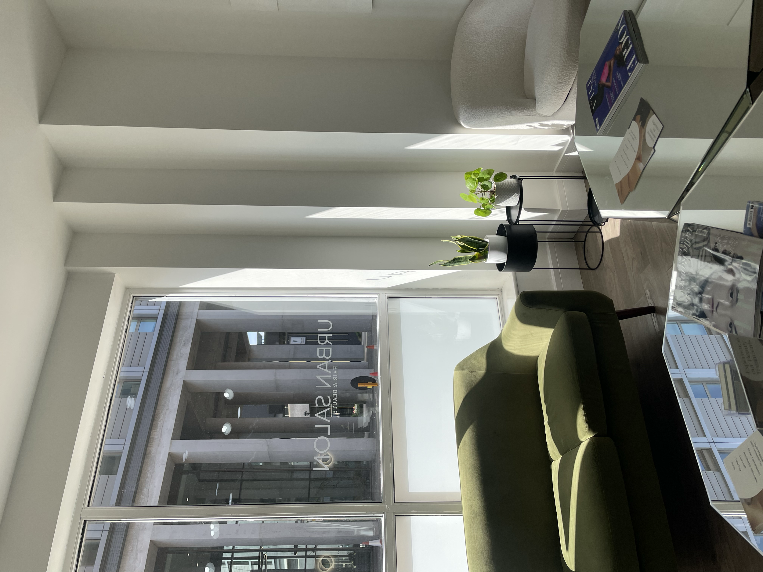 Modern office waiting area with a green sofa, two potted plants, a glass table with magazines, and a large window with a view of the building exterior.