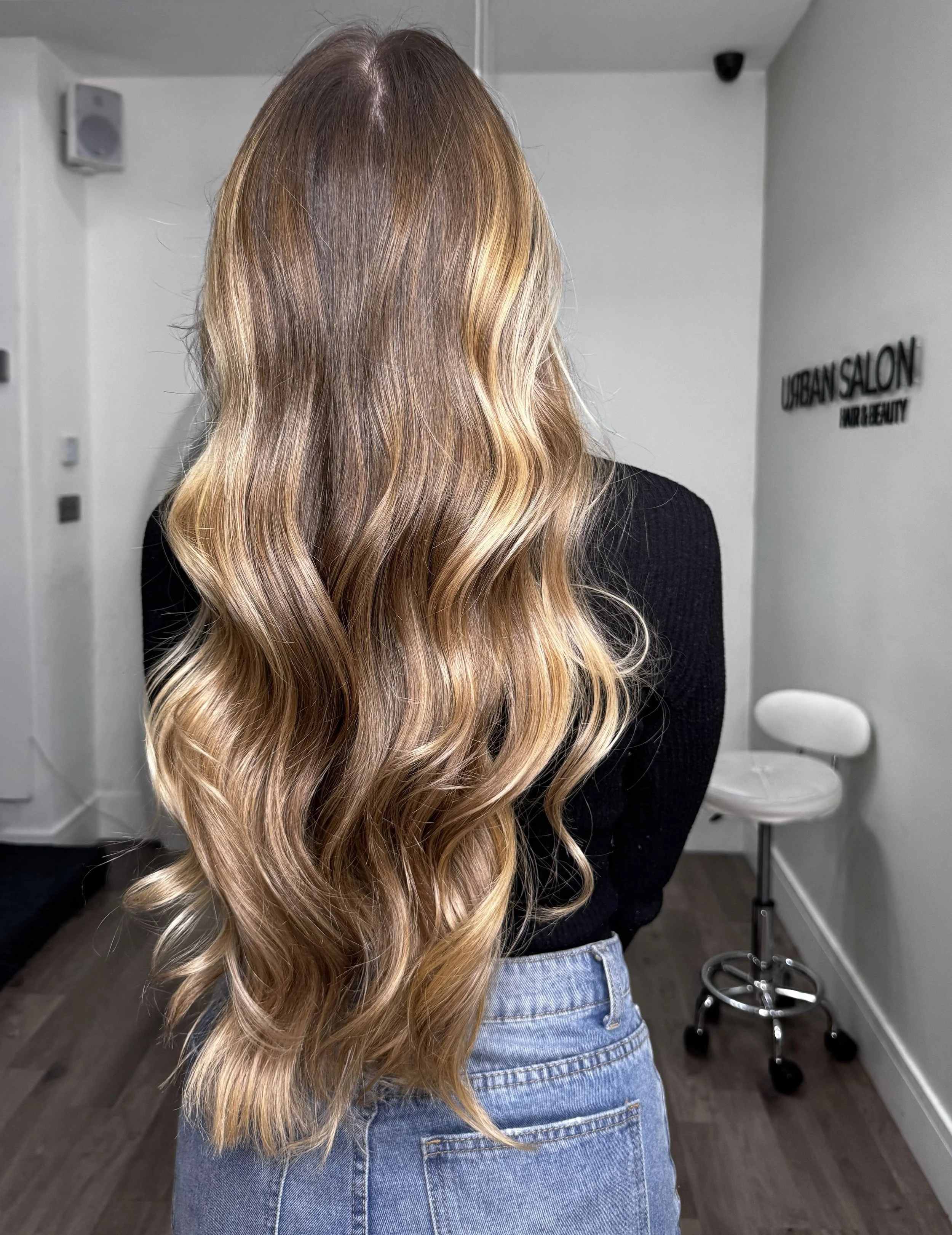 Back view of a woman with long, wavy, blonde hair in a salon.
