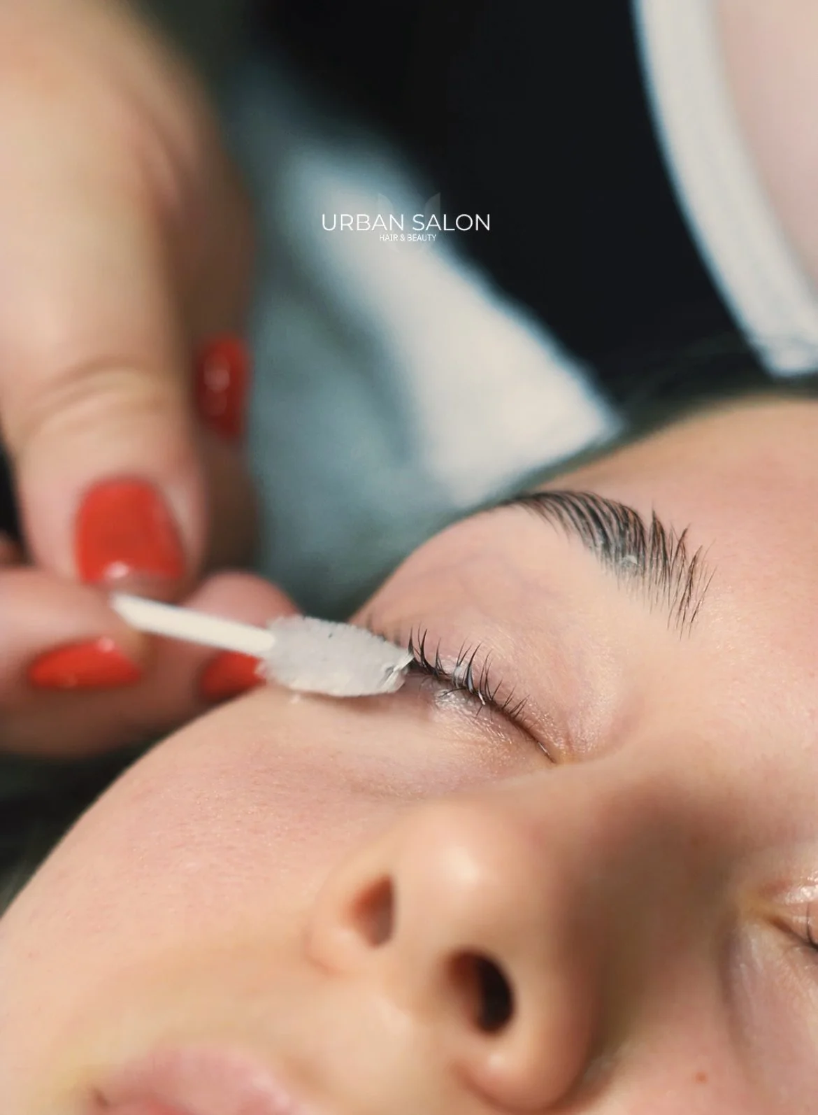 Close-up of a woman receiving eyelash extension treatment at an urban salon, with an esthetician applying adhesive to her eyelashes using a cotton swab.