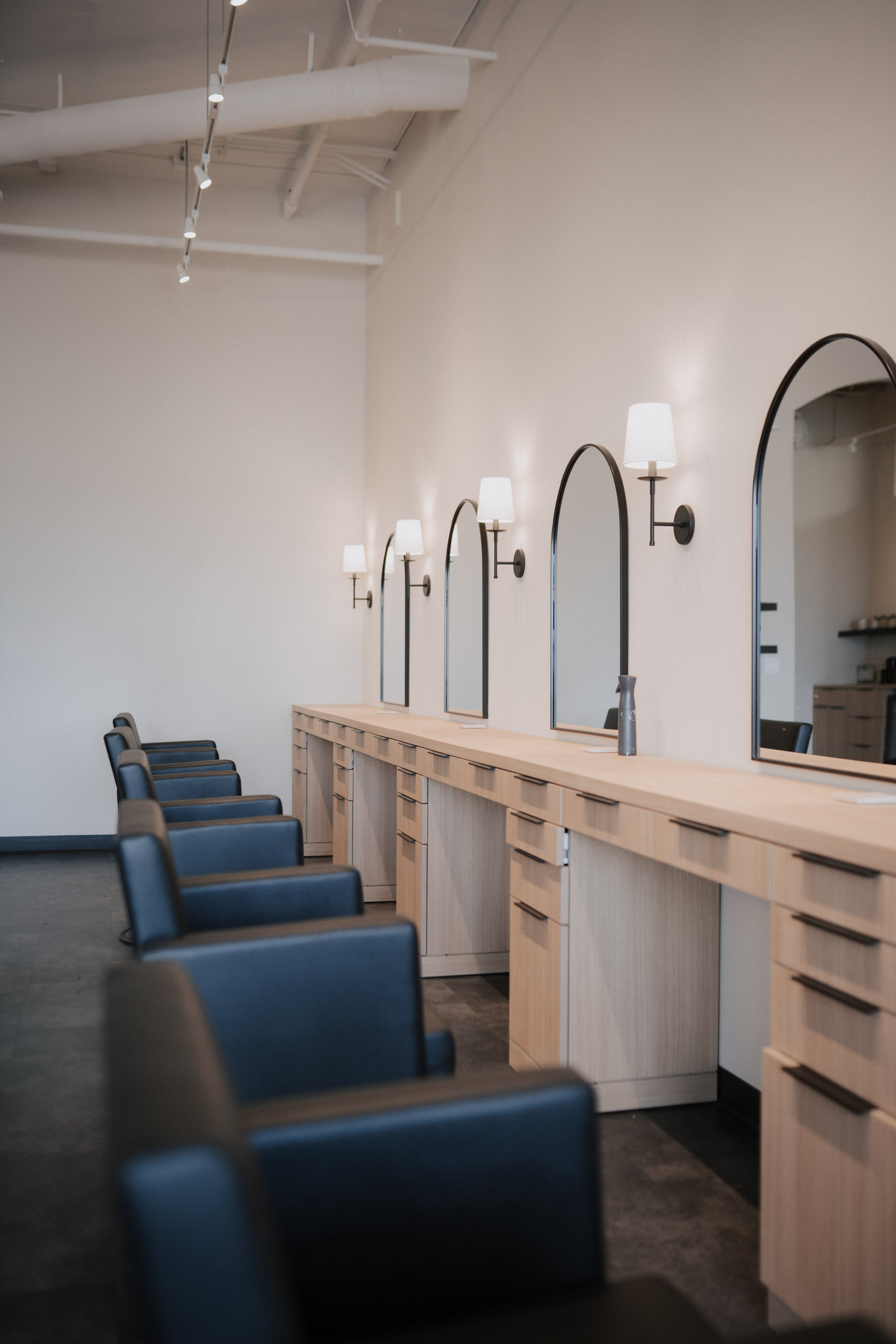 Empty salon with black chairs, light wood stations, large mirrors, and wall-mounted lamps in a modern, minimalistic style.