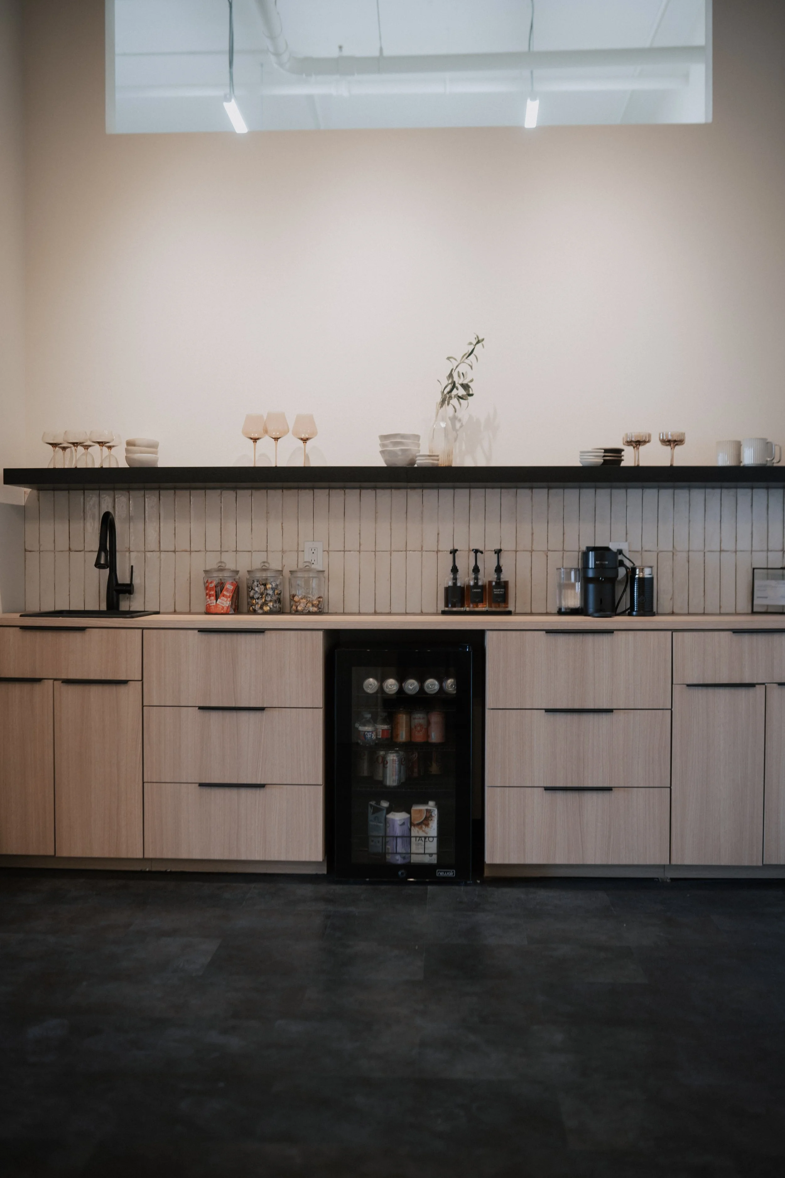 Minimalist kitchen with light wood cabinets, black countertop, and a small beverage fridge, decorated with glassware and bowls.
