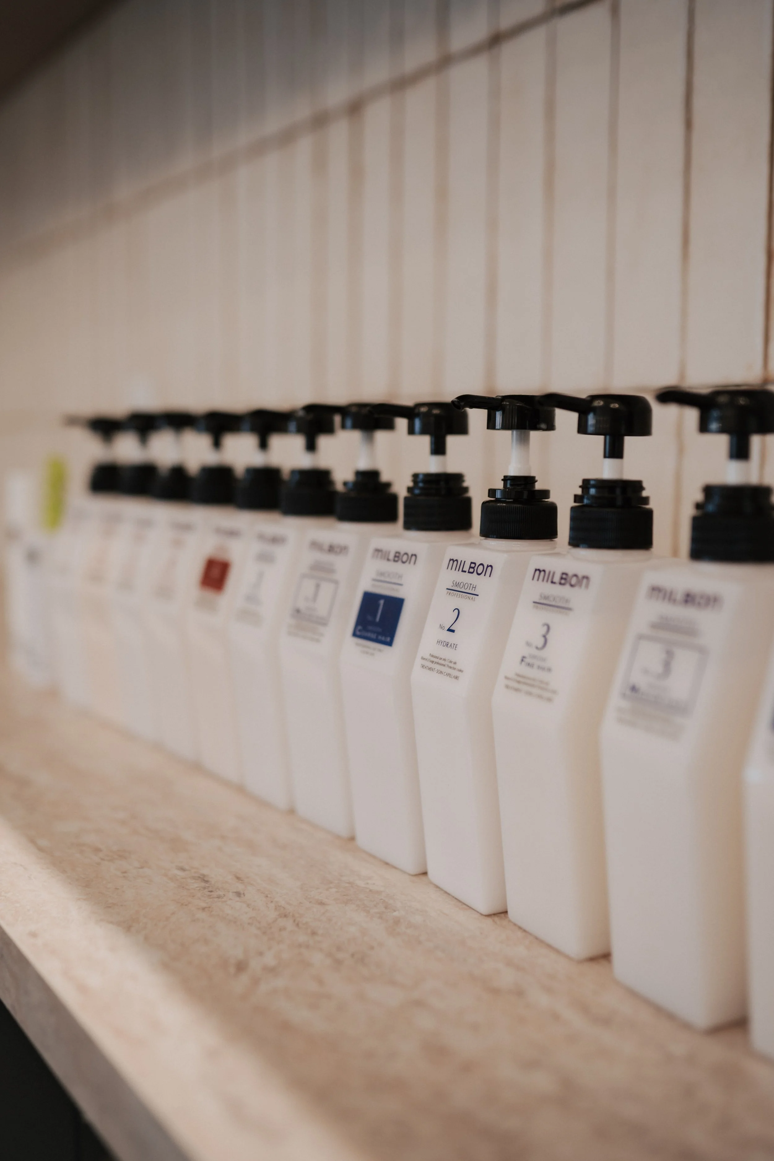 A row of white bottles with black pump tops, lined up on a wooden surface, labeled with different numbers and colors, likely in a salon or spa setting.