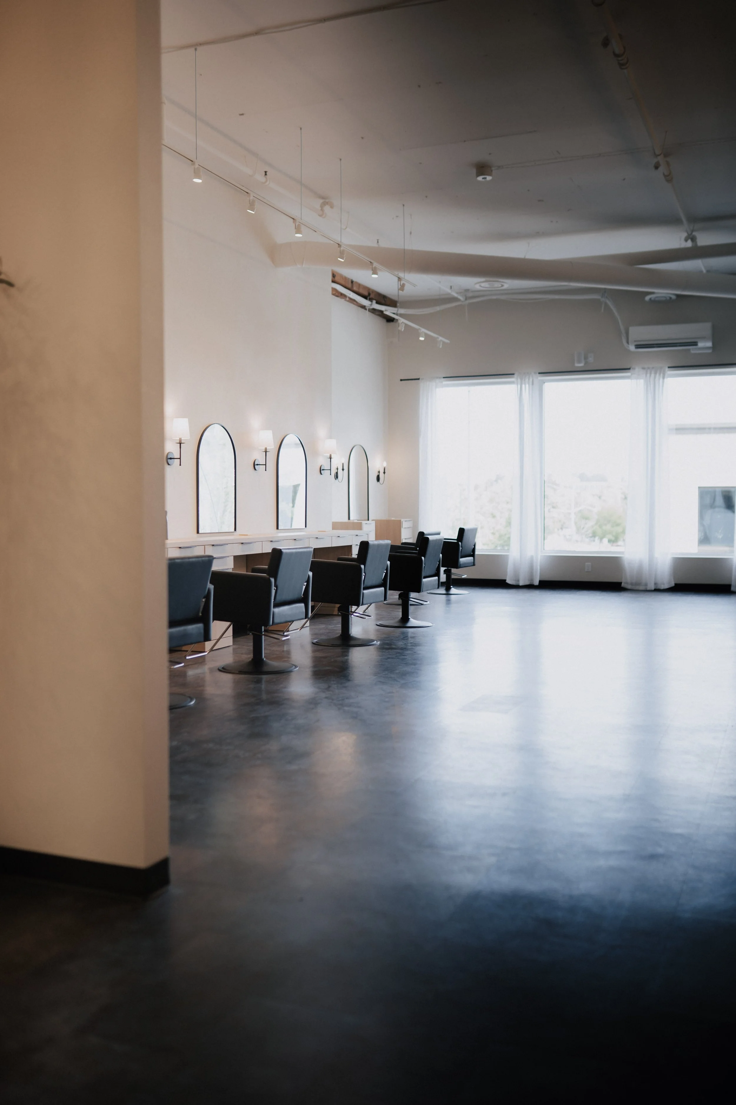 Empty hair salon with black styling chairs, mirrors, and large windows with white curtains.