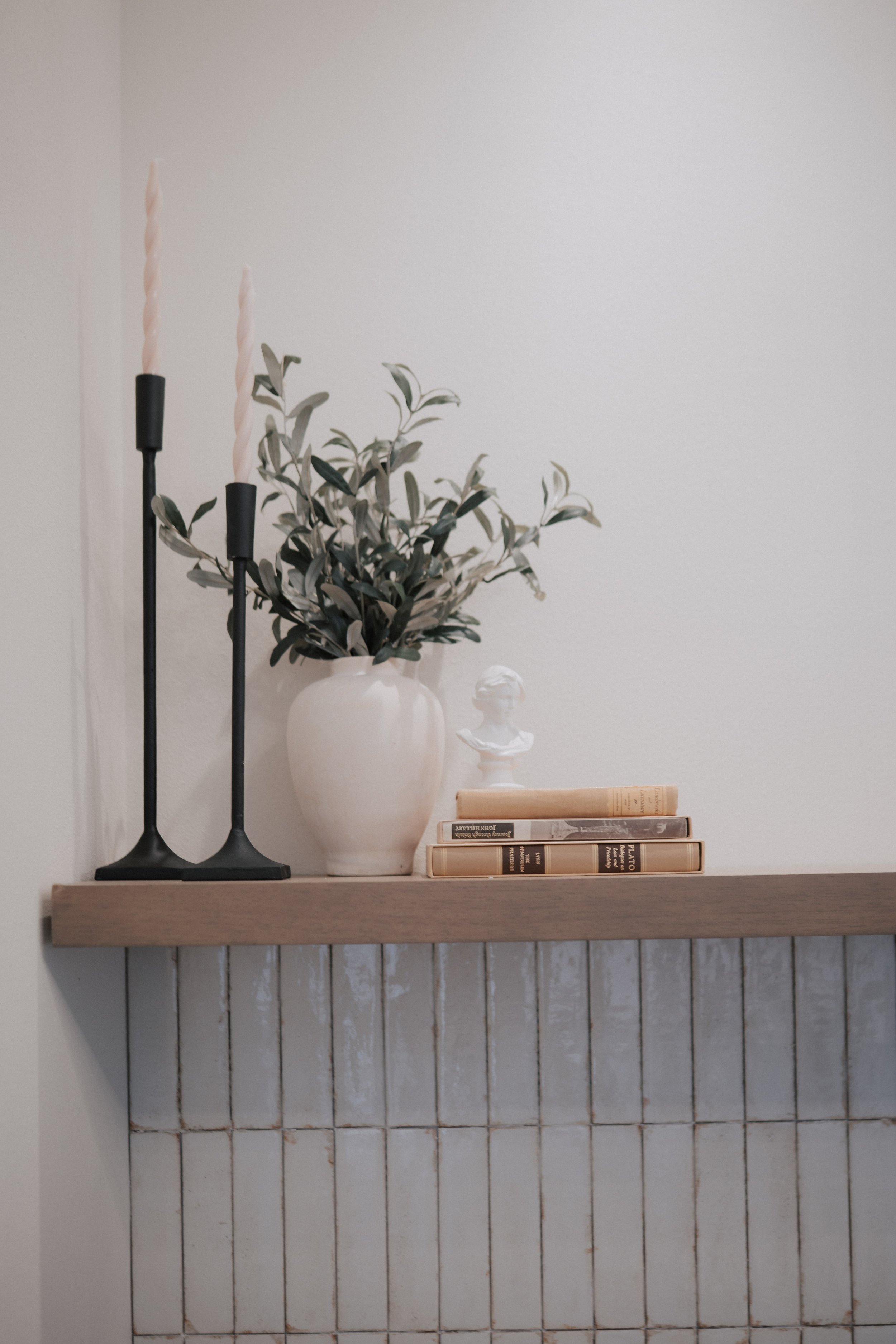 Decorative mantel with three pink candles on black candleholders, a white vase with green leafy plant, small bust sculpture, and a stack of three books against a plain wall.