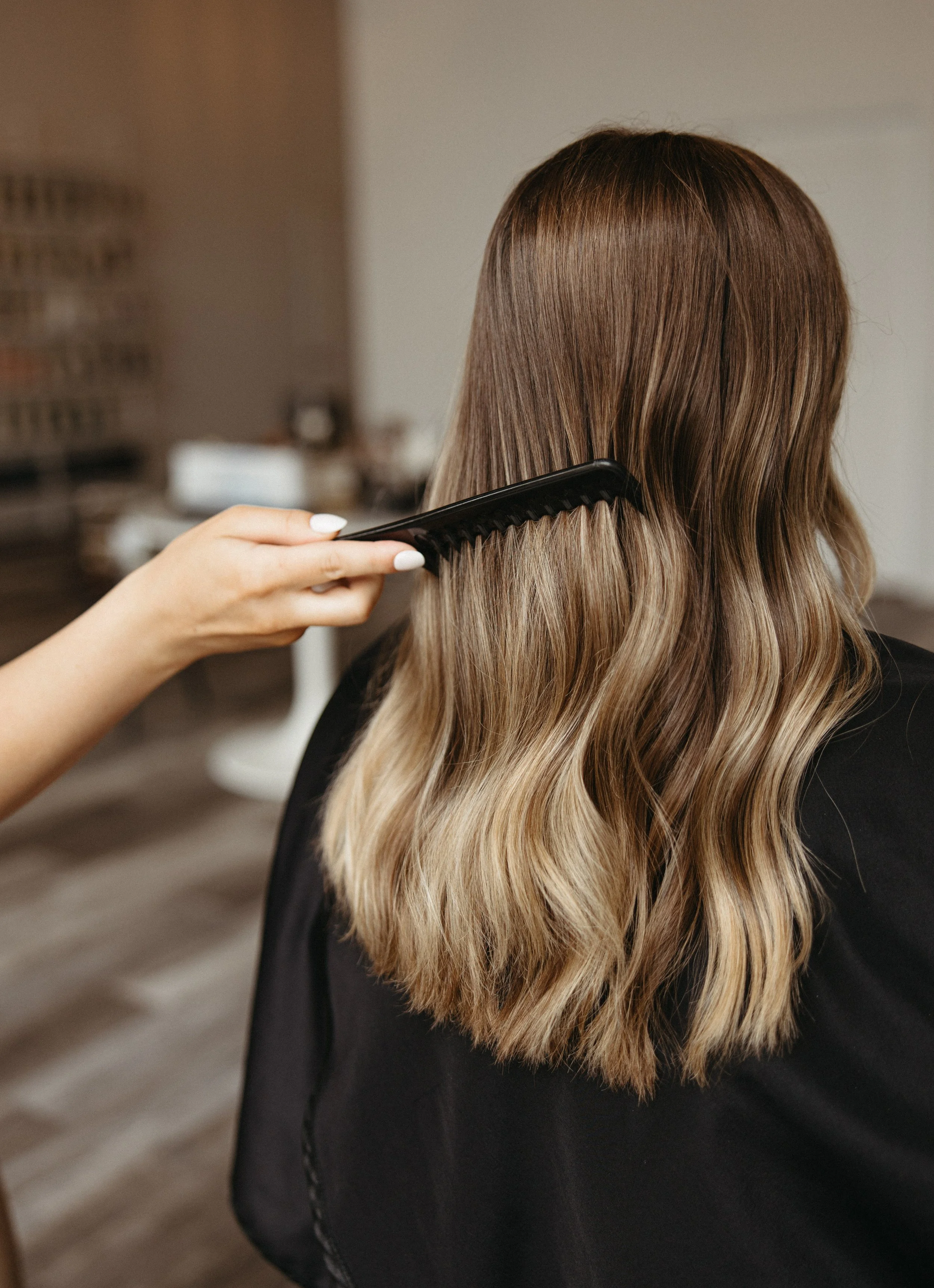 A person with long, wavy, light brown hair is being combed by a stylist in a salon.