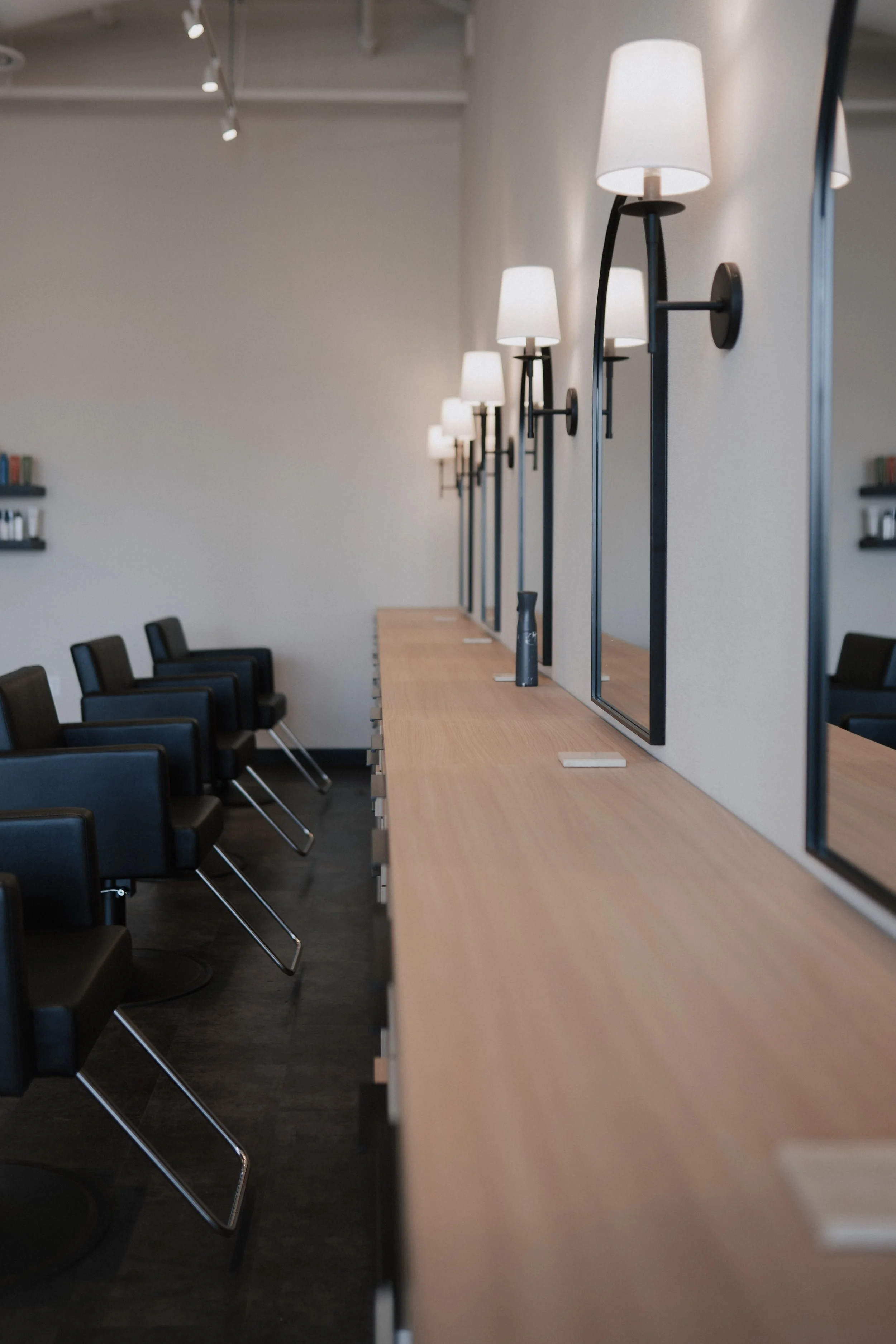 Empty salon with black chairs, wooden counter, wall-mounted lamps, and mirrors.