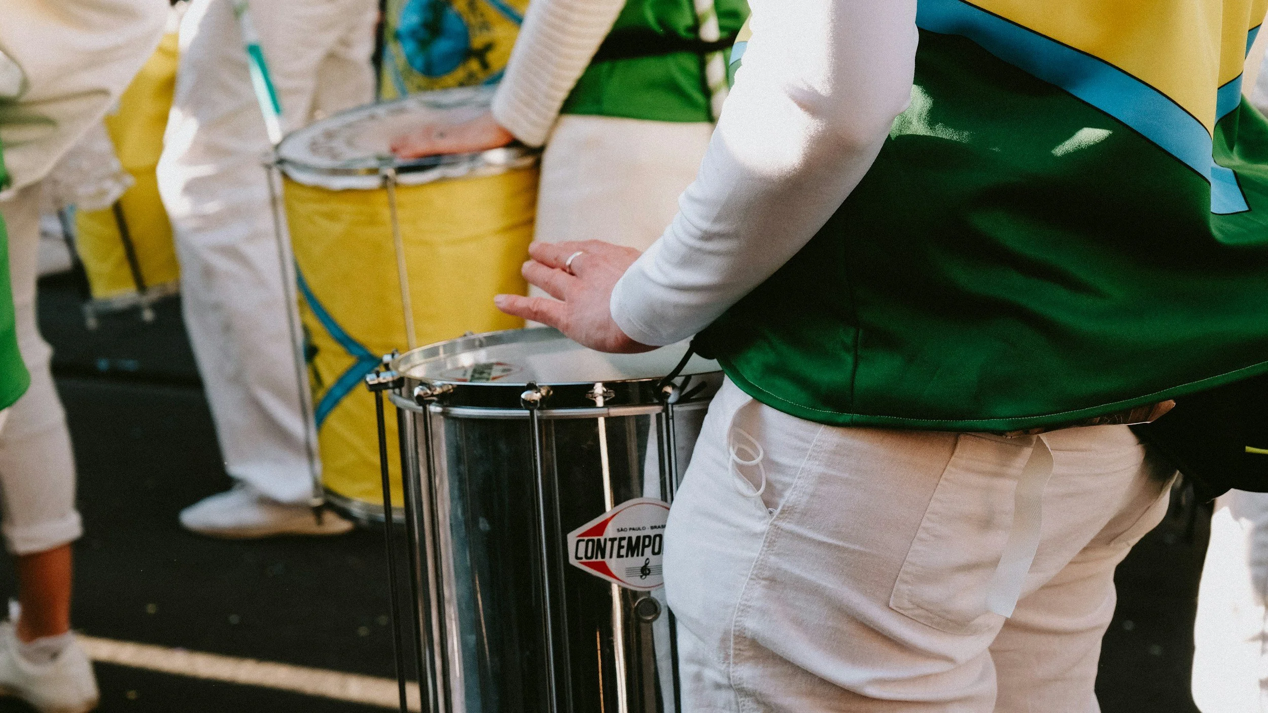 Person with a white long sleeve shirt and beige pants playing a drum, surrounded by other drummers with yellow and white drums in an outdoor setting.