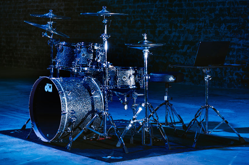 A silver drum set with cymbals, a laptop on a stand, and a stool, in a dimly lit room with a brick wall background.