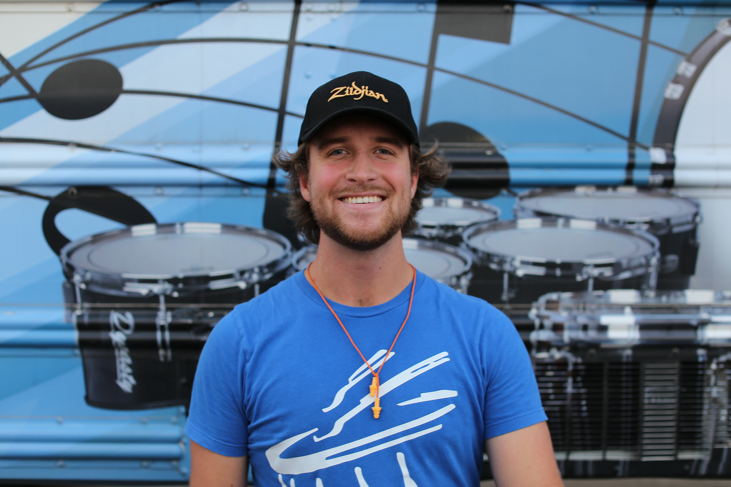A smiling young man with a beard and brown hair, wearing a black cap and blue t-shirt, standing in front of a black, white, and blue mural featuring large illustrations of drums.