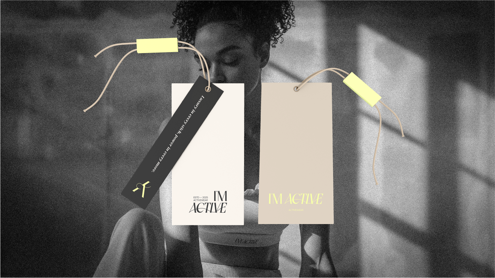 Black and white photo of a woman with curly hair looking down, wearing an activewear top. In the foreground, there are three hanging tags or labels with the text 'I'M ACTIVE' and related branding.