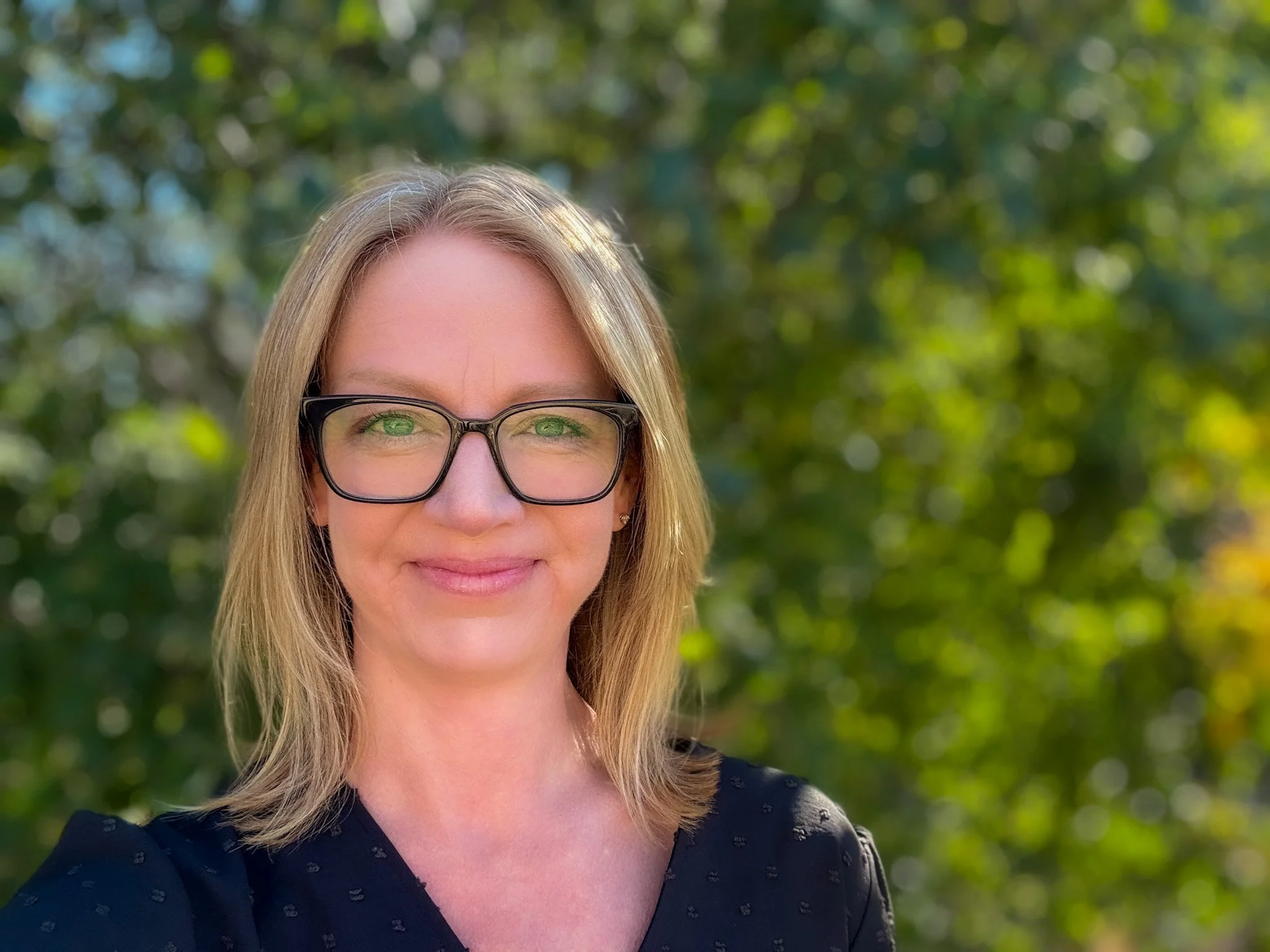 A woman with blonde hair and glasses smiling outdoors with a blurred background of green trees.