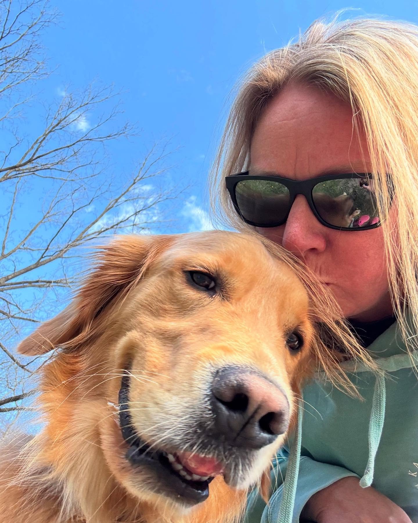 A woman wearing sunglasses taking a selfie with her golden retriever dog outdoors under a blue sky with a few wispy clouds and bare tree branches in the background.
