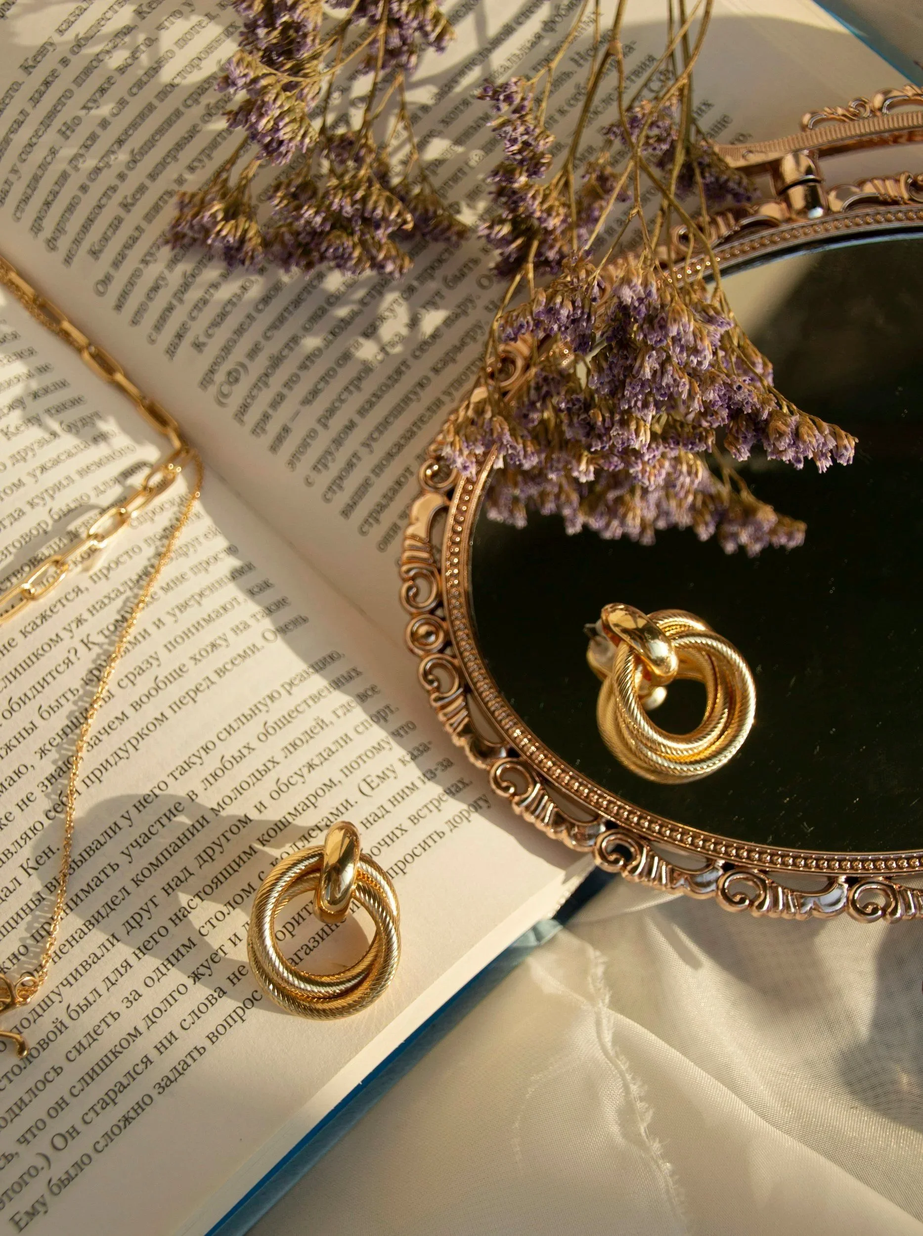 Gold jewelry, including a ring and a necklace, on an open book, with dried purple flowers and a vintage mirror with a decorative frame.