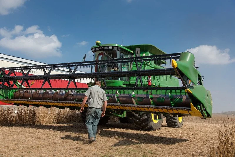 A man walking towards a large green and yellow combine harvester in a field under a blue sky with scattered clouds.