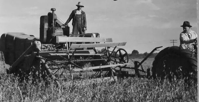Black and white photo of three farmers working in a field with a tractor and vintage farming equipment.