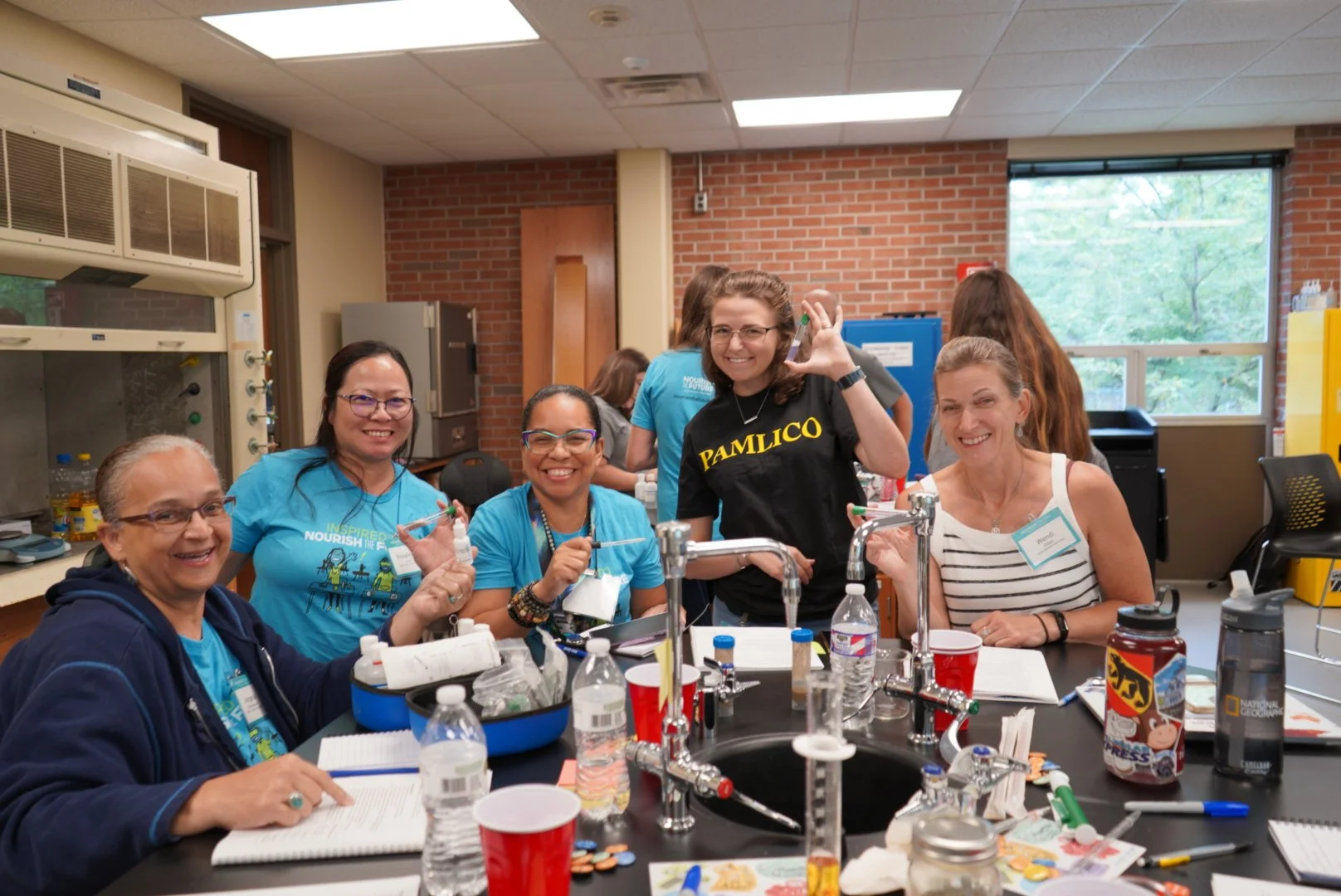 A group of six women smiling and posing for the camera in a science classroom or lab, with lab equipment on the table, including water bottles, notebooks, and supplies. The background shows a brick wall, windows, and laboratory furniture.