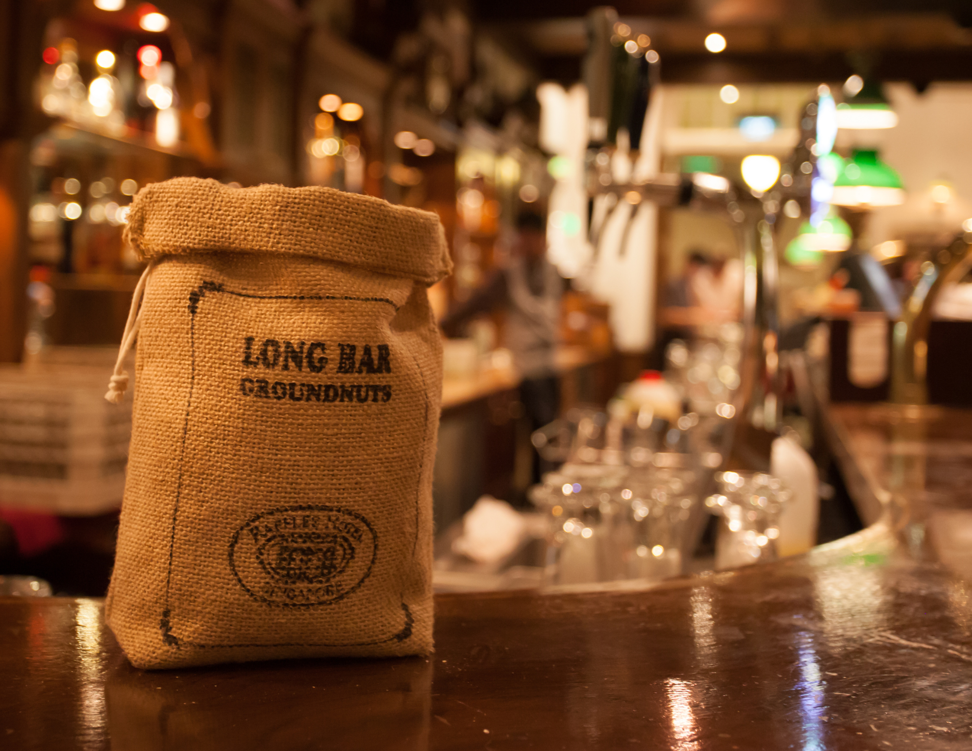 A burlap sack labeled "Long Bar Groundnuts" sitting on a bar counter with a blurred bar background, including glasses, bottles, and patrons.