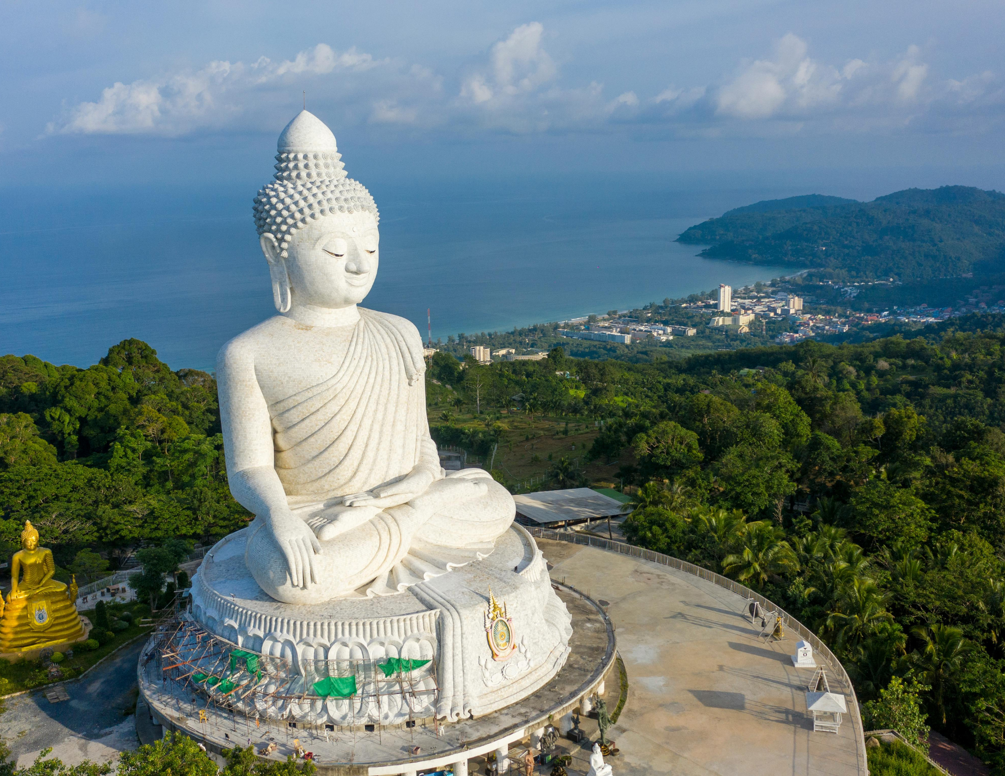 A large white Buddha statue in a seated meditation pose with a serene expression, overlooking a coastal town with lush greenery and mountains in the background.