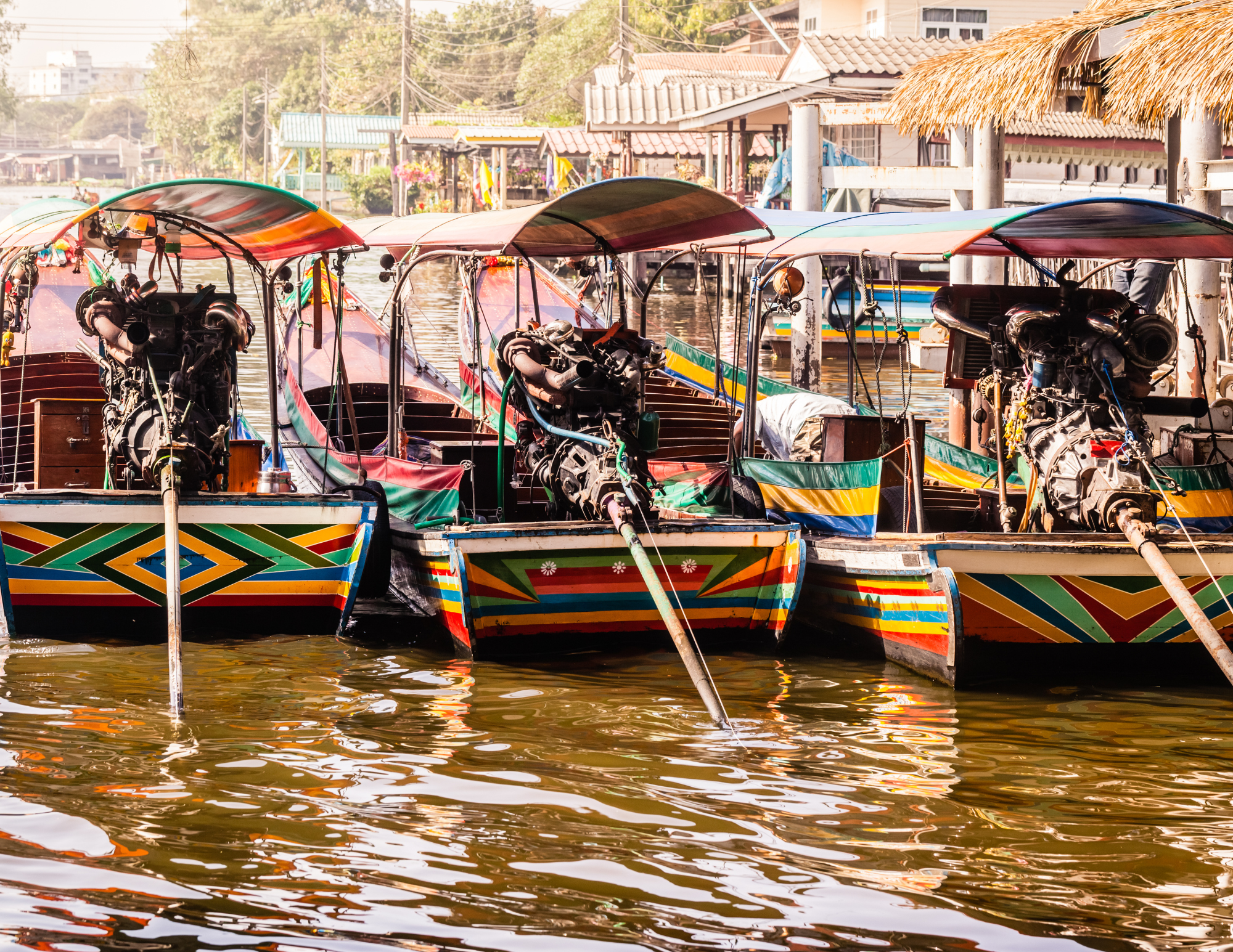 Several colorful boats with outboard motors docked at a riverside with buildings and huts in the background.