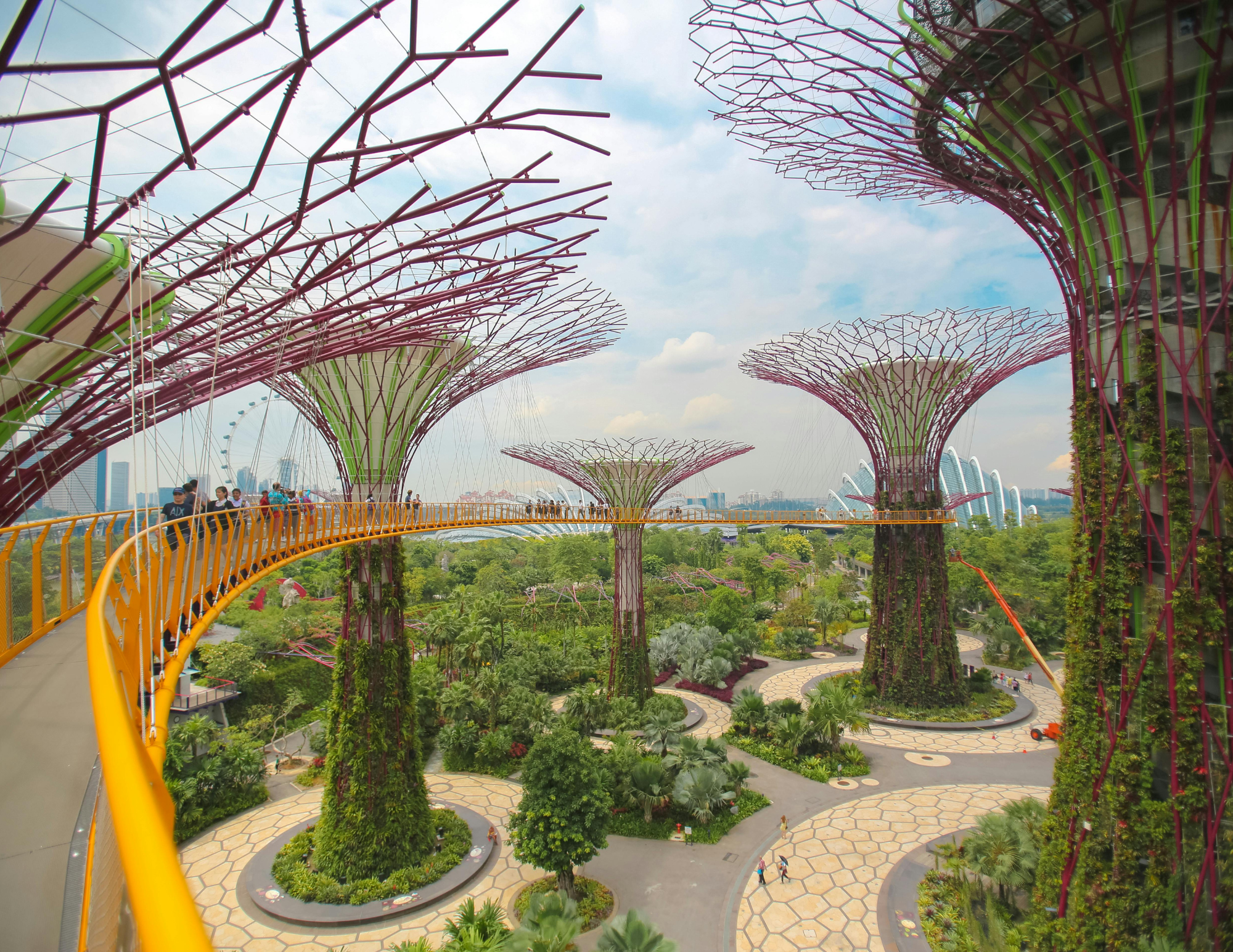People walking on the Orange Bridge in Gardens by the Bay, Singapore, with futuristic Supertree Grove structures and lush greenery below.