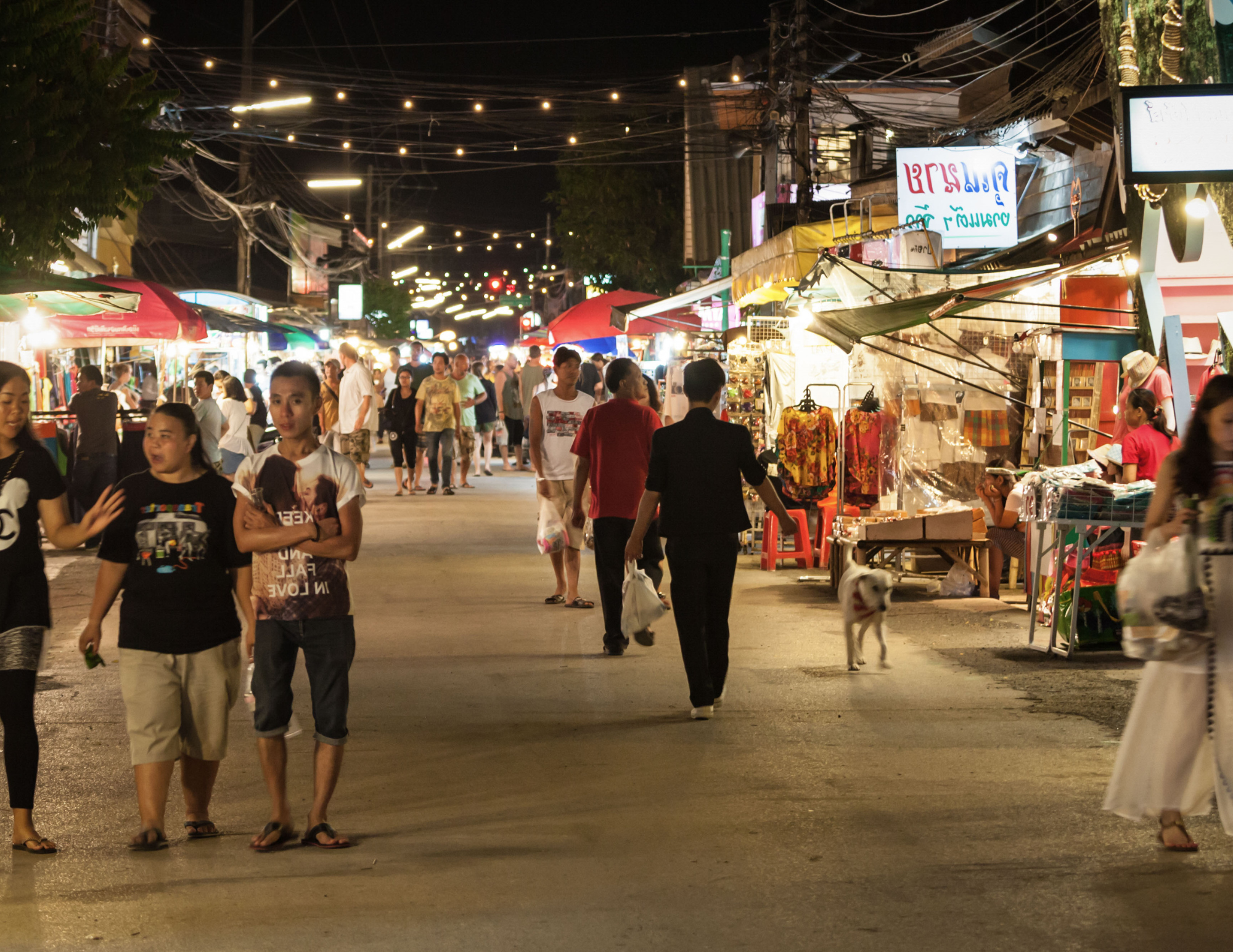 People walking along a bustling street market at night, illuminated by string lights and shop signs, with stalls selling clothing and accessories.