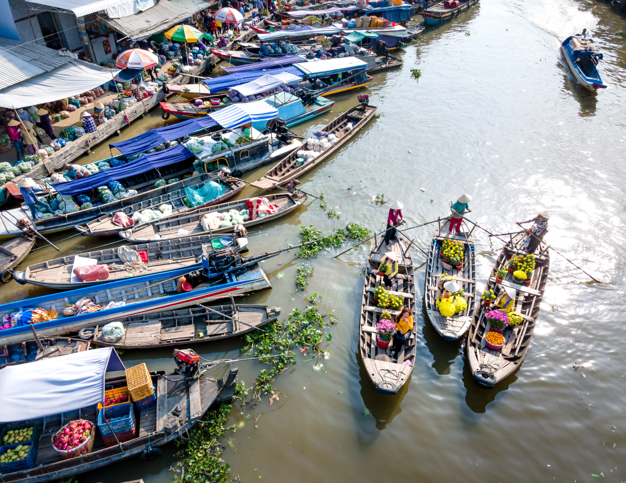 Boats docked along a riverbank at a floating market, with vendors selling flowers, fruits, and other goods on the boats and on the dock, and vendors wearing traditional conical hats.