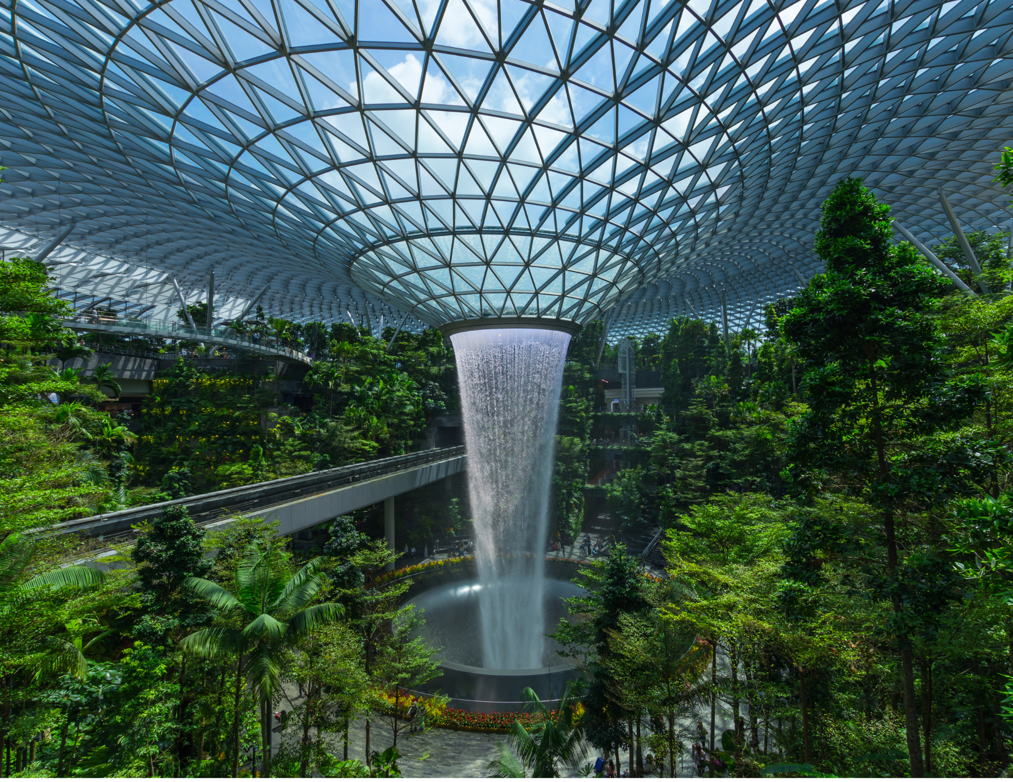 Indoor garden with a large waterfall falling from a glass-domed ceiling into a pool below, surrounded by lush green trees and plants.