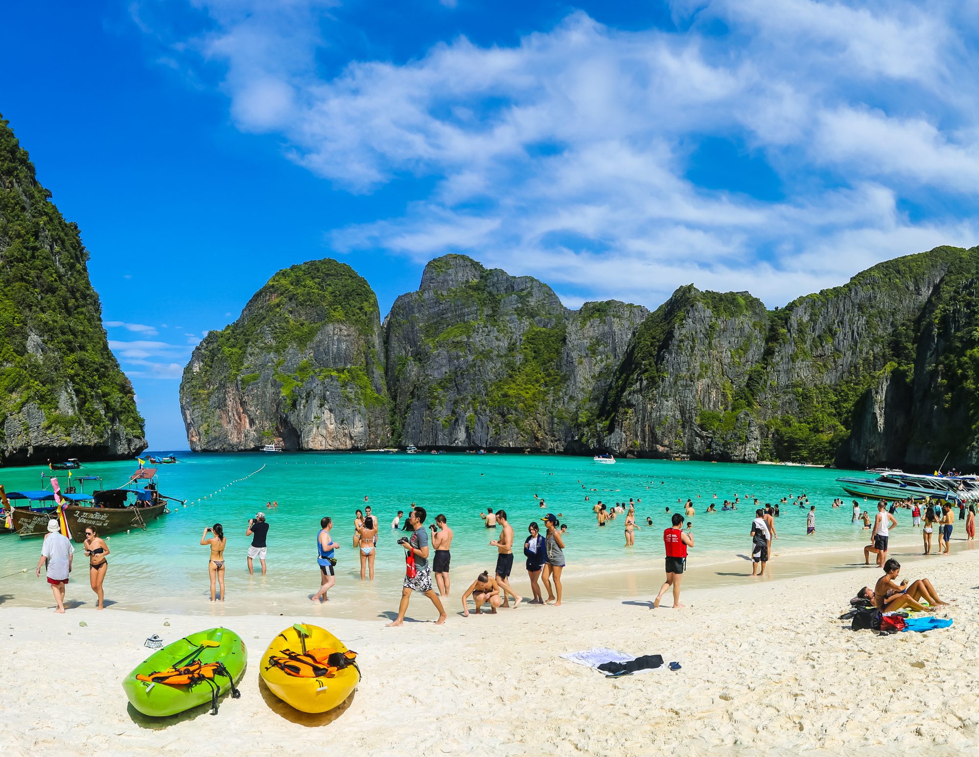 People enjoying a sunny day at a tropical beach with turquoise water, surrounded by cliffs and lush green mountains, some swimming, some relaxing on the sand, kayaks in the foreground, boats docked nearby.