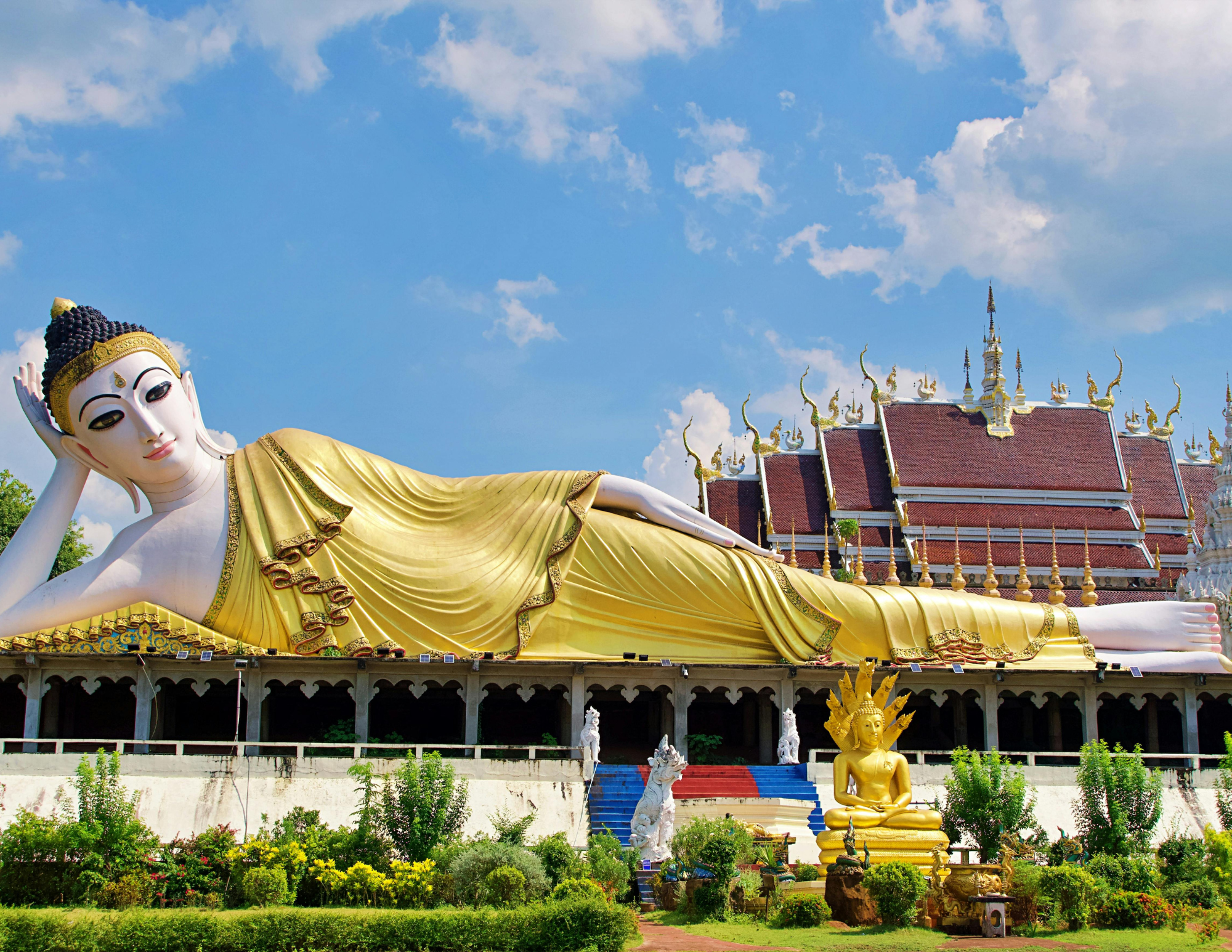 Large reclining Buddha statue with a gold robe, set against a sky with clouds, with traditional Thai architecture and smaller statues in the surroundings.
