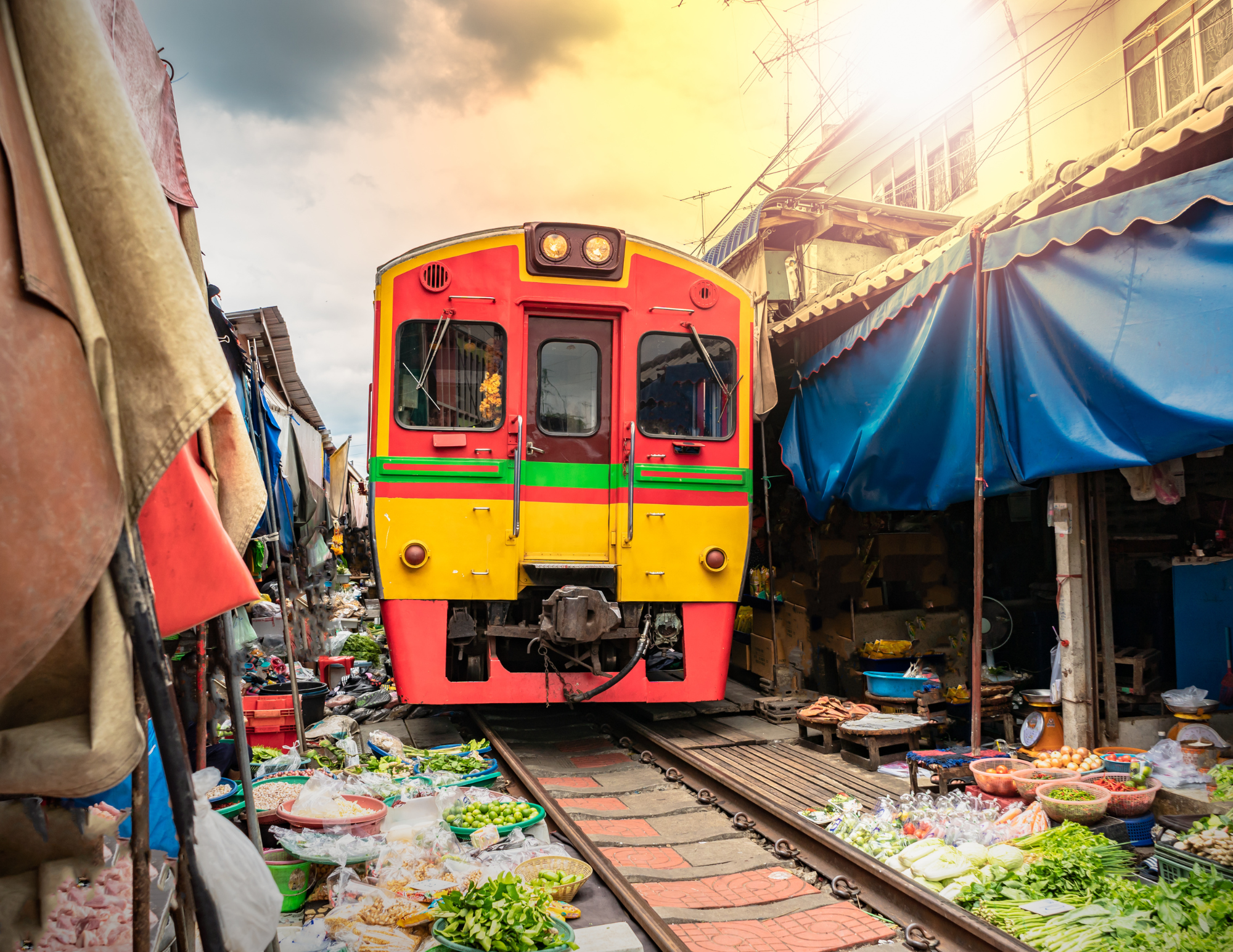 A colorful train is approaching through a marketplace with vegetable stalls on both sides, covered by awnings and tarps.