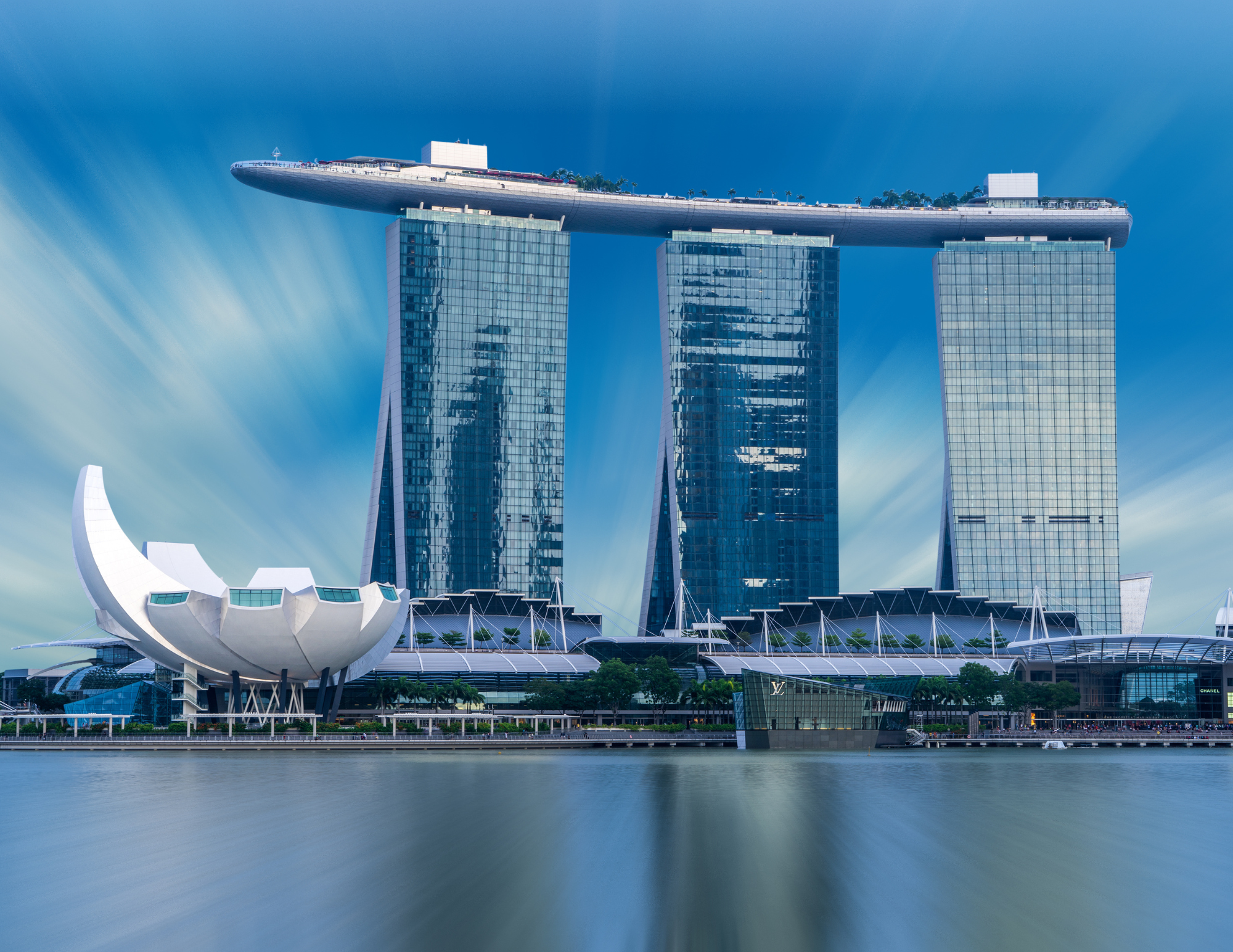Modern cityscape featuring the Marina Bay Sands hotel in Singapore with three skyscraper towers and a boat-shaped structure on top, alongside other contemporary buildings and a waterfront.