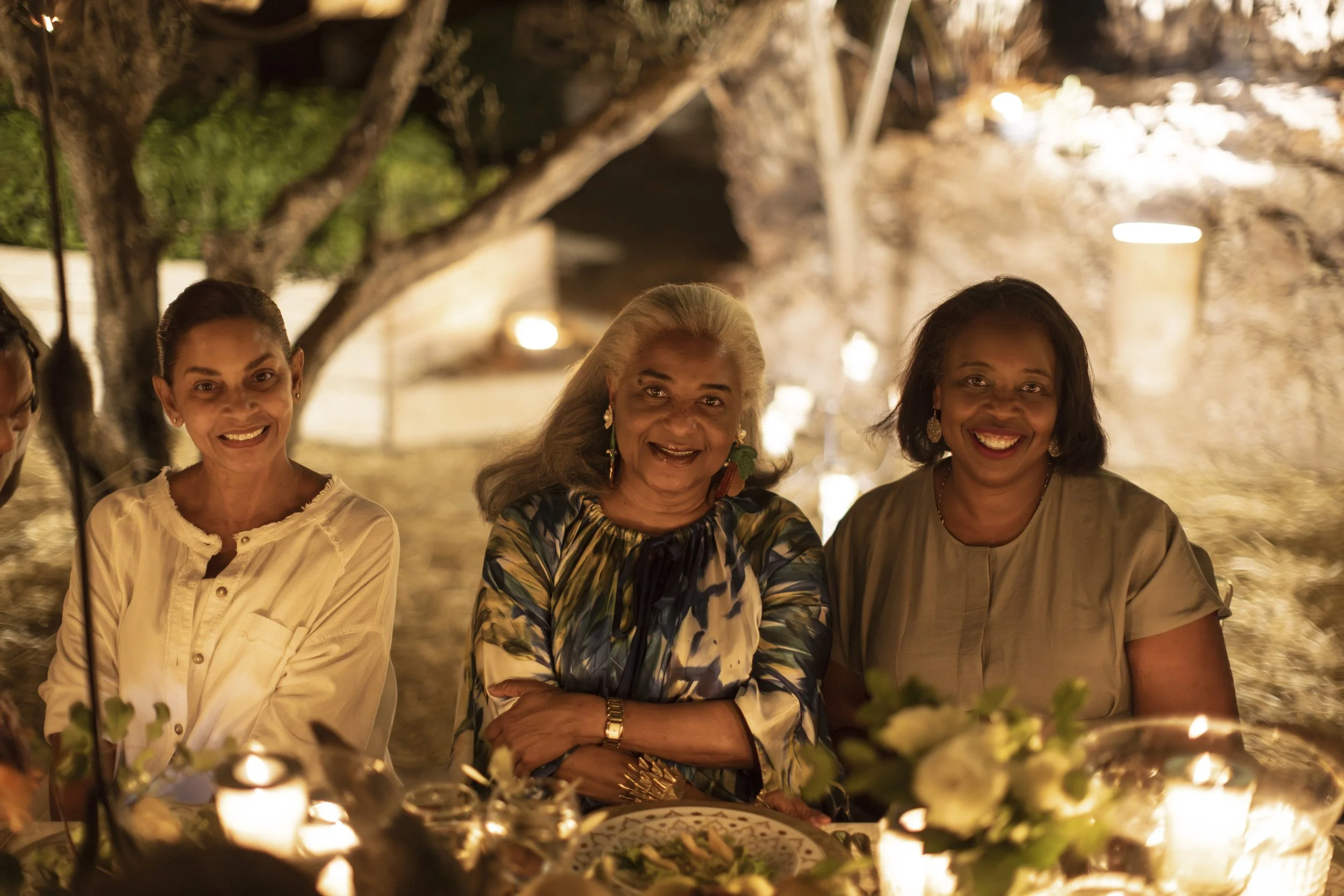 Three women smiling and sitting at a dinner party, in an outdoor setting with warm lighting and trees in the background.