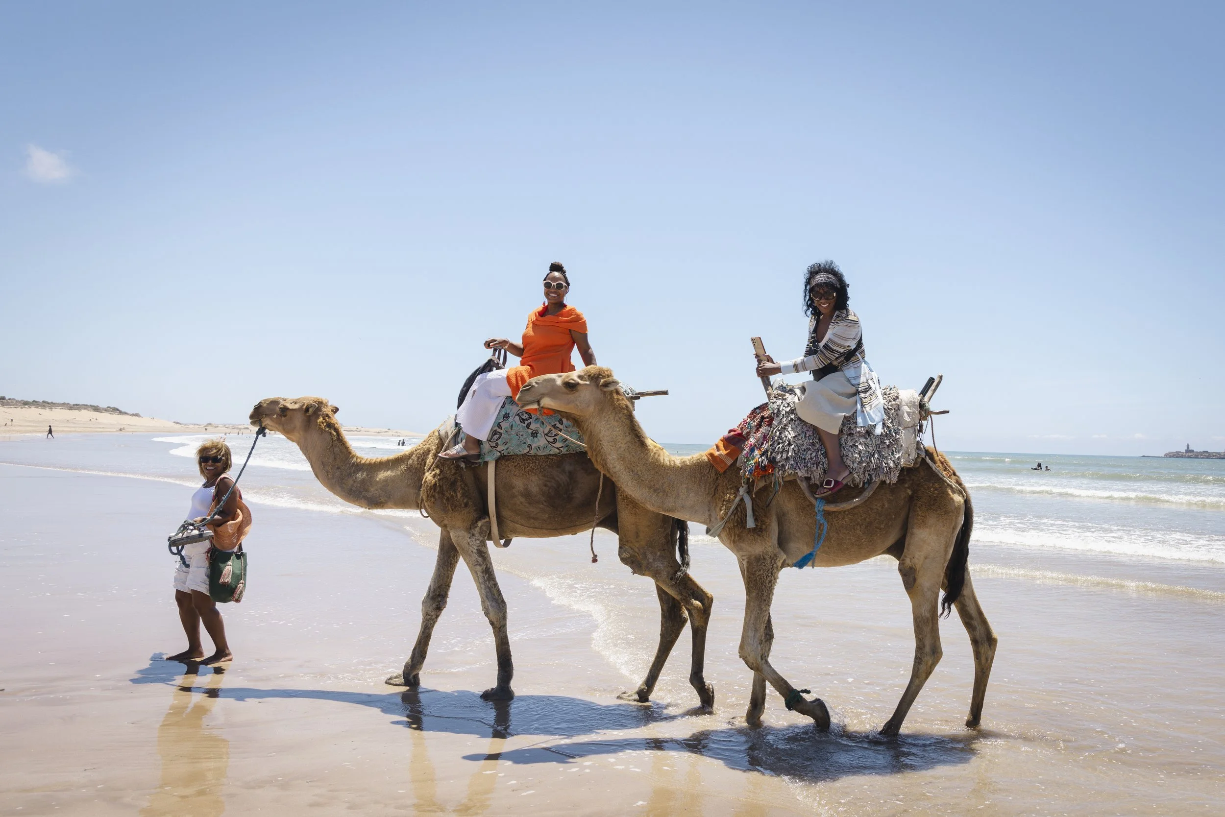 Three women and two camels on the beach, with the ocean in the background. Two women are riding camels, and one woman is walking beside the camels, holding a leash.