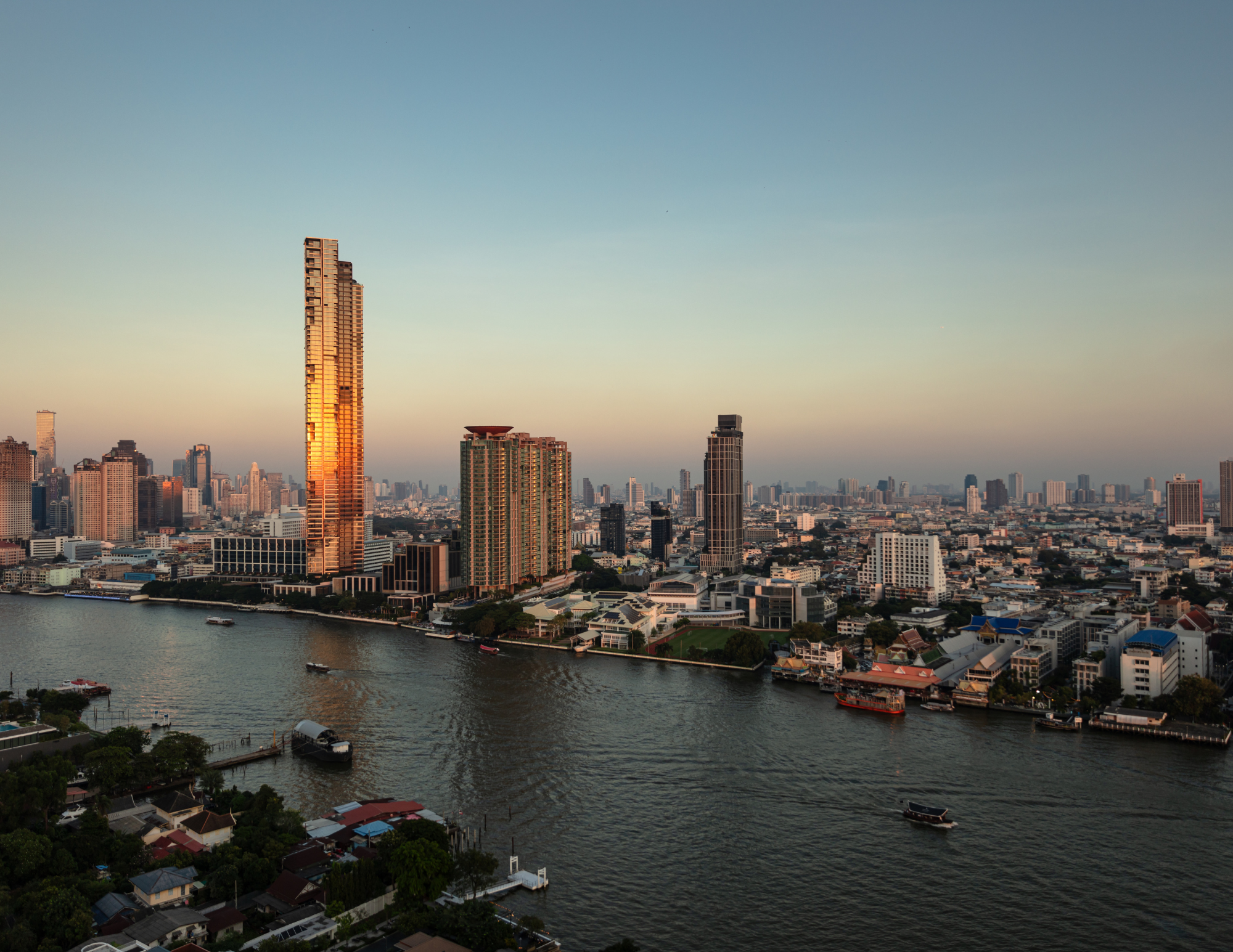 City skyline at sunset with tall buildings and a river in the foreground, boats on the water, and a clear sky.