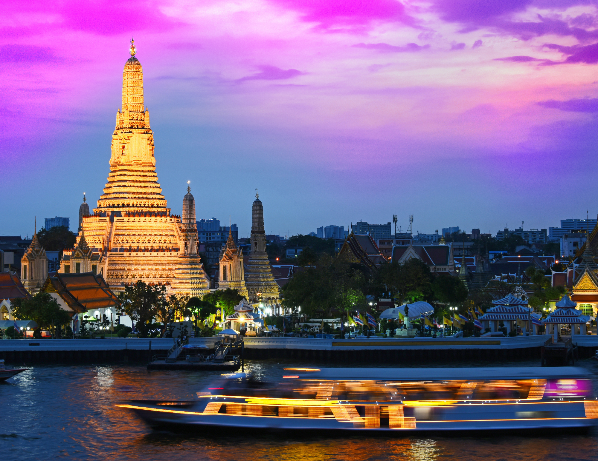 A golden illuminated temple with a tiered spire on a river, surrounded by smaller temples and buildings, with a purple and pink sky at sunset and a boat passing on the water.