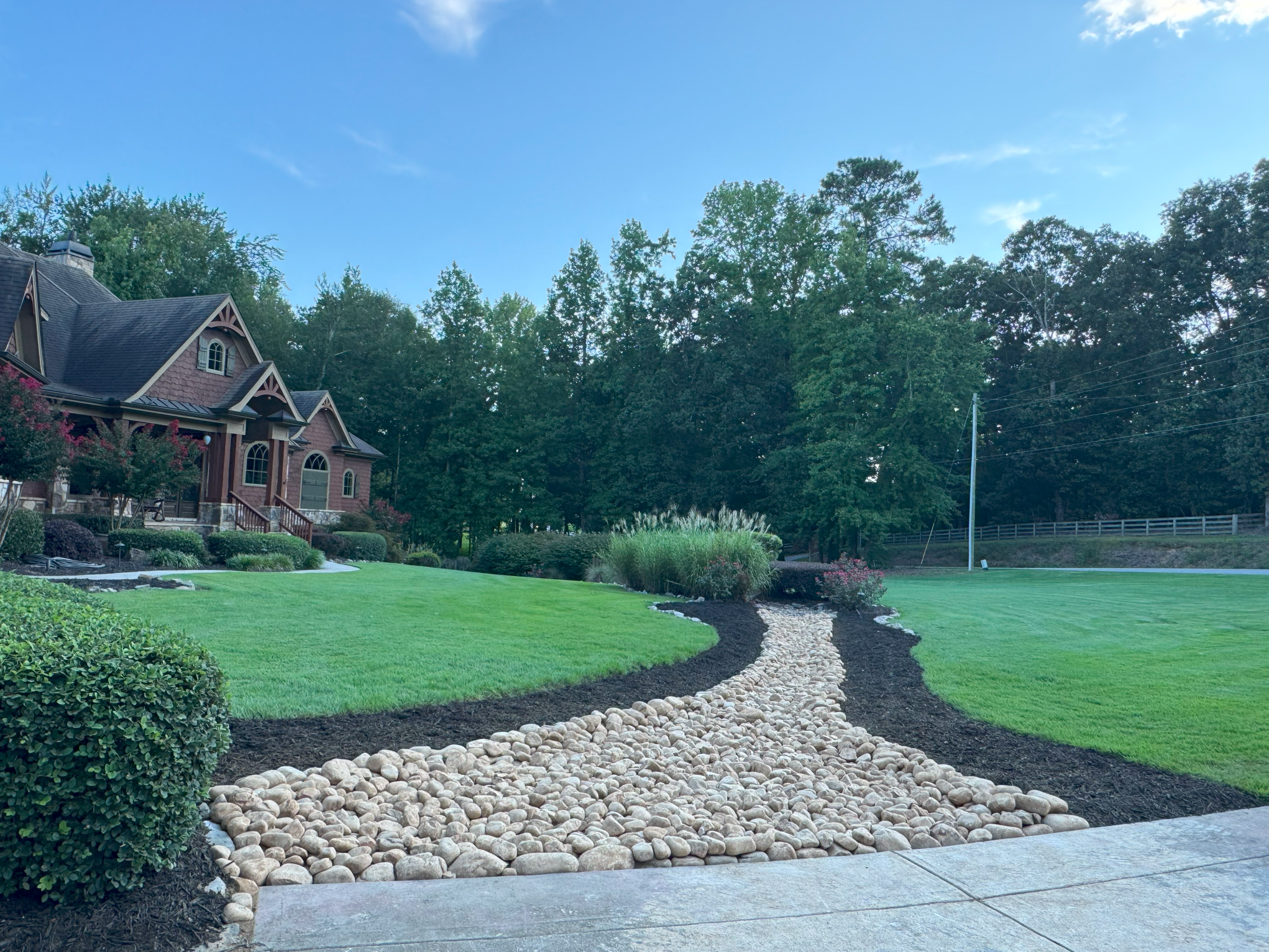 A landscaped yard with a curved pebble path, green grass, bushes, and trees, in front of a house with a steep roof and arched windows under a blue sky.