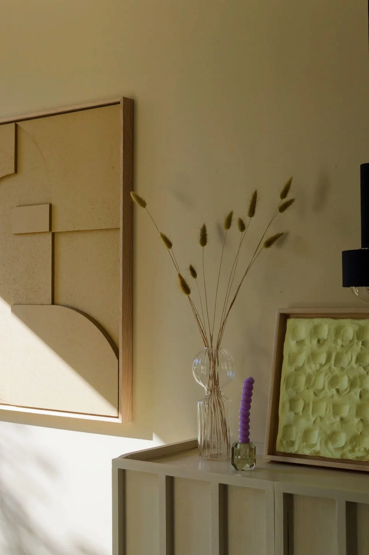 Decorative interior with a clear glass vase holding dried grass, a purple candle in a small glass holder, and abstract art pieces on a light-colored wooden cabinet.