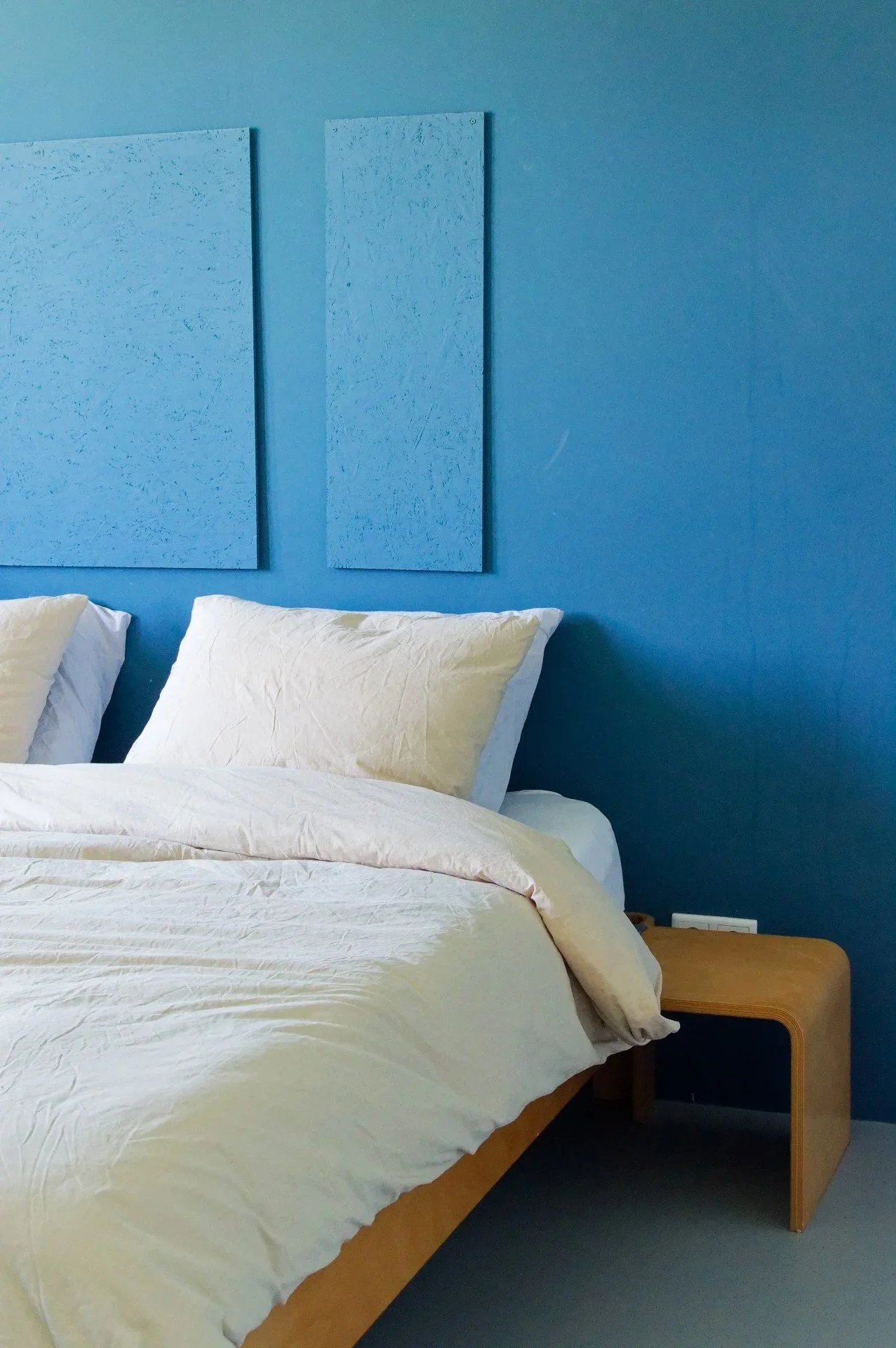 A bedroom with a blue wall, two light-colored pillows, and a bed with white bedding, next to a wooden nightstand.