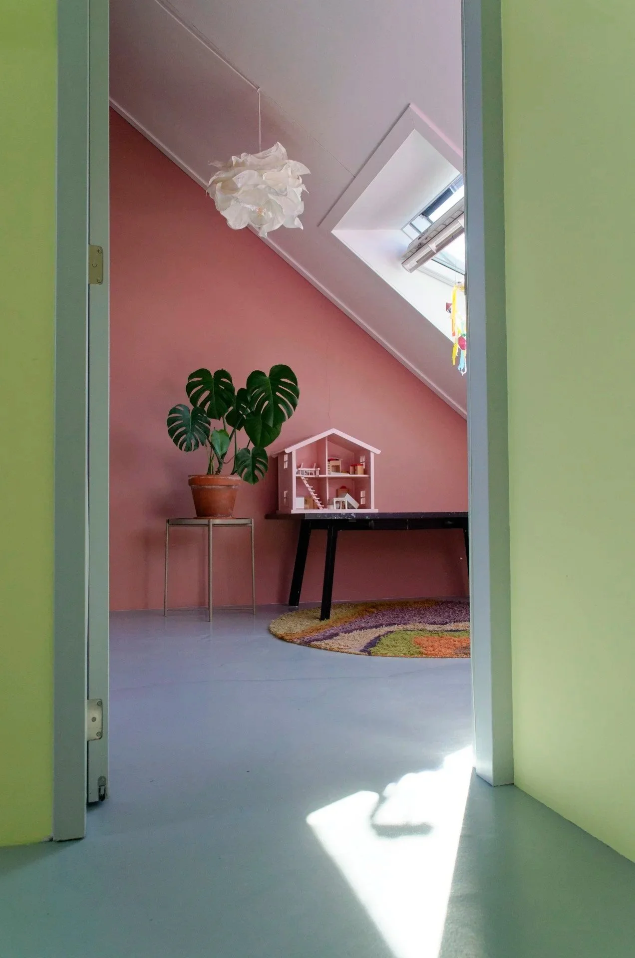 View of a cozy indoor room seen through a doorway, featuring a pink wall, a large green plant in a pot on a stand, a small dollhouse, a black table, a colorful rug, and a skylight window with sunlight streaming in.