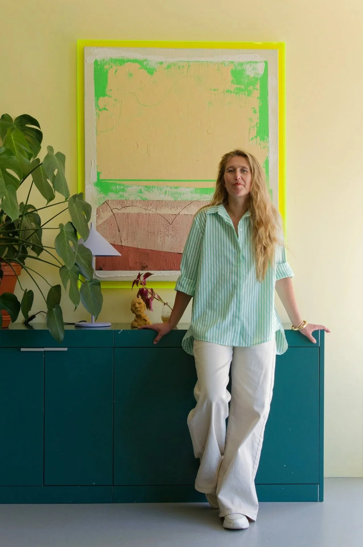 A woman with long blonde hair in a green and white striped shirt and white pants standing in front of a teal cabinet, with a large leafy plant on the left and abstract artwork on the wall behind her.