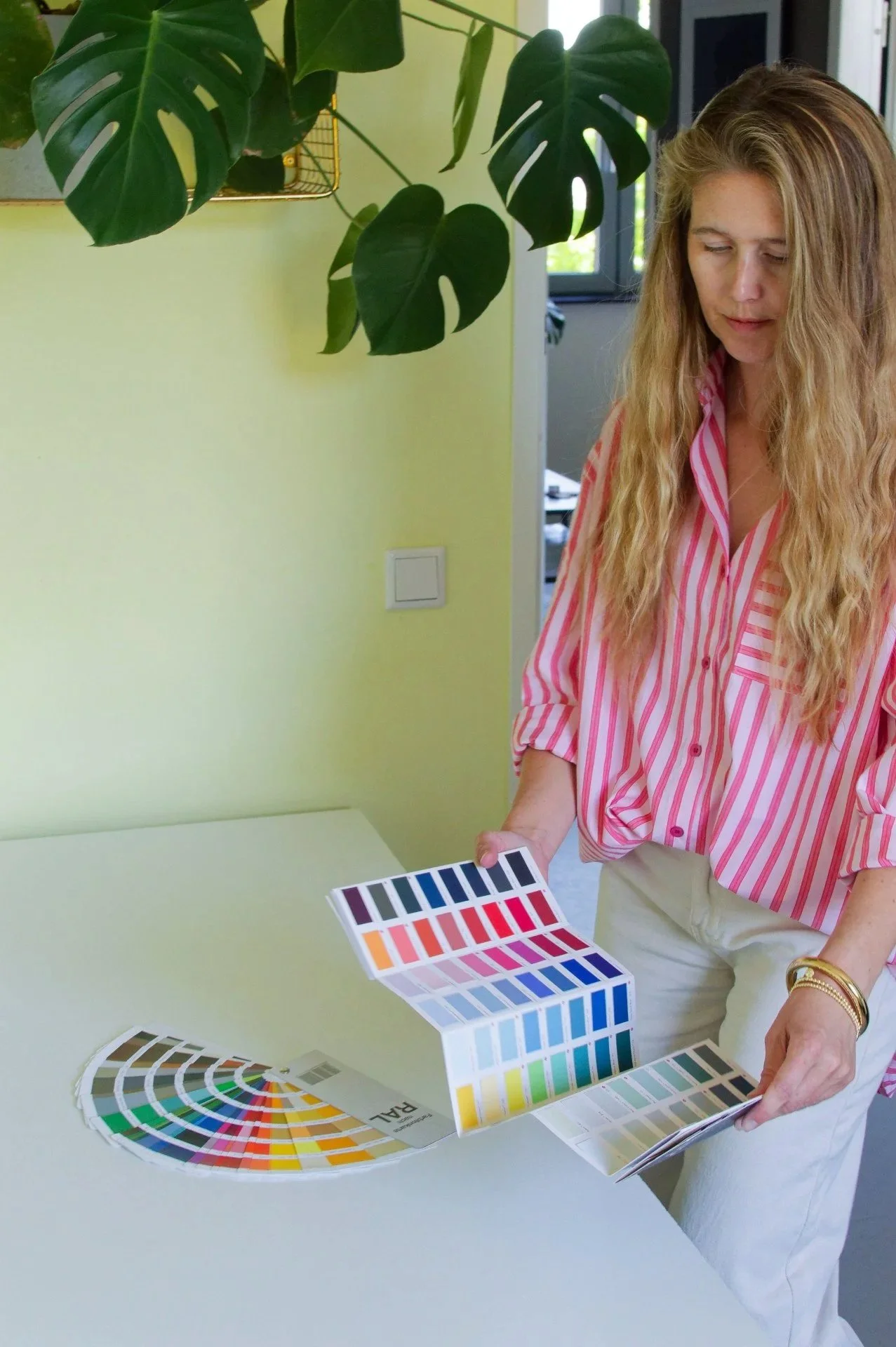 Woman with long blonde hair in a pink and white striped shirt looking at color swatches on a white table in a room with green plants and a yellow wall.