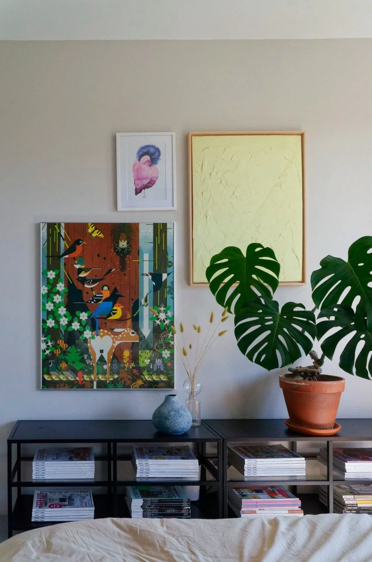 Decorative interior wall with framed artwork, potted Monstera plant, and magazines on black shelves.