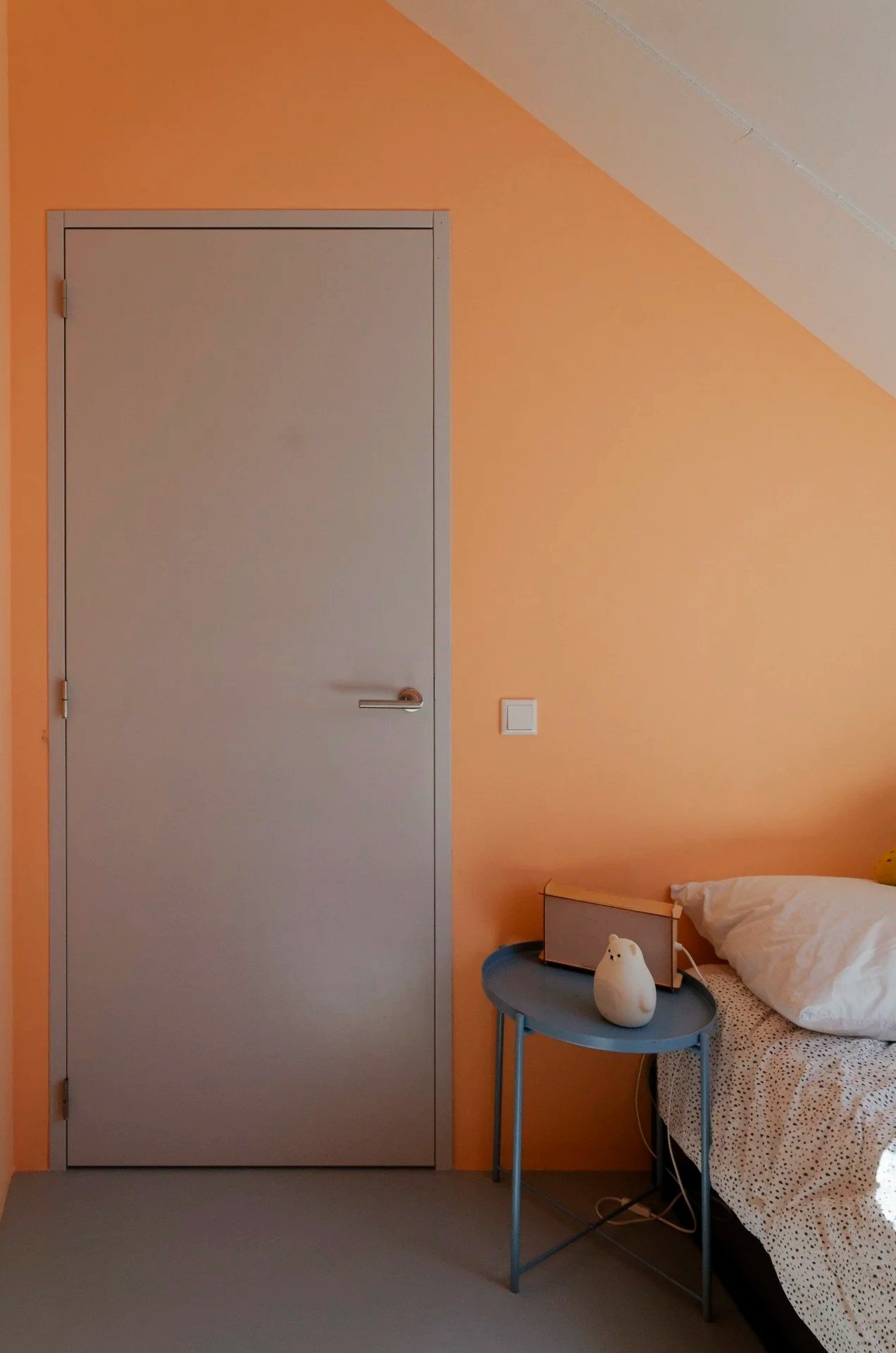 A bedroom corner with a closed gray door, an orange wall, a small blue side table with a white decorative object, a bed with white and dotted sheets, a book, and a soft white pillow.