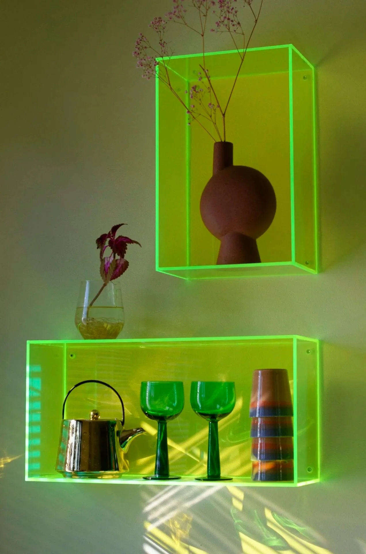 Neon green illuminated wall shelves holding glassware, a teapot, a vase with flowers, and a ceramic vase with dried flowers.