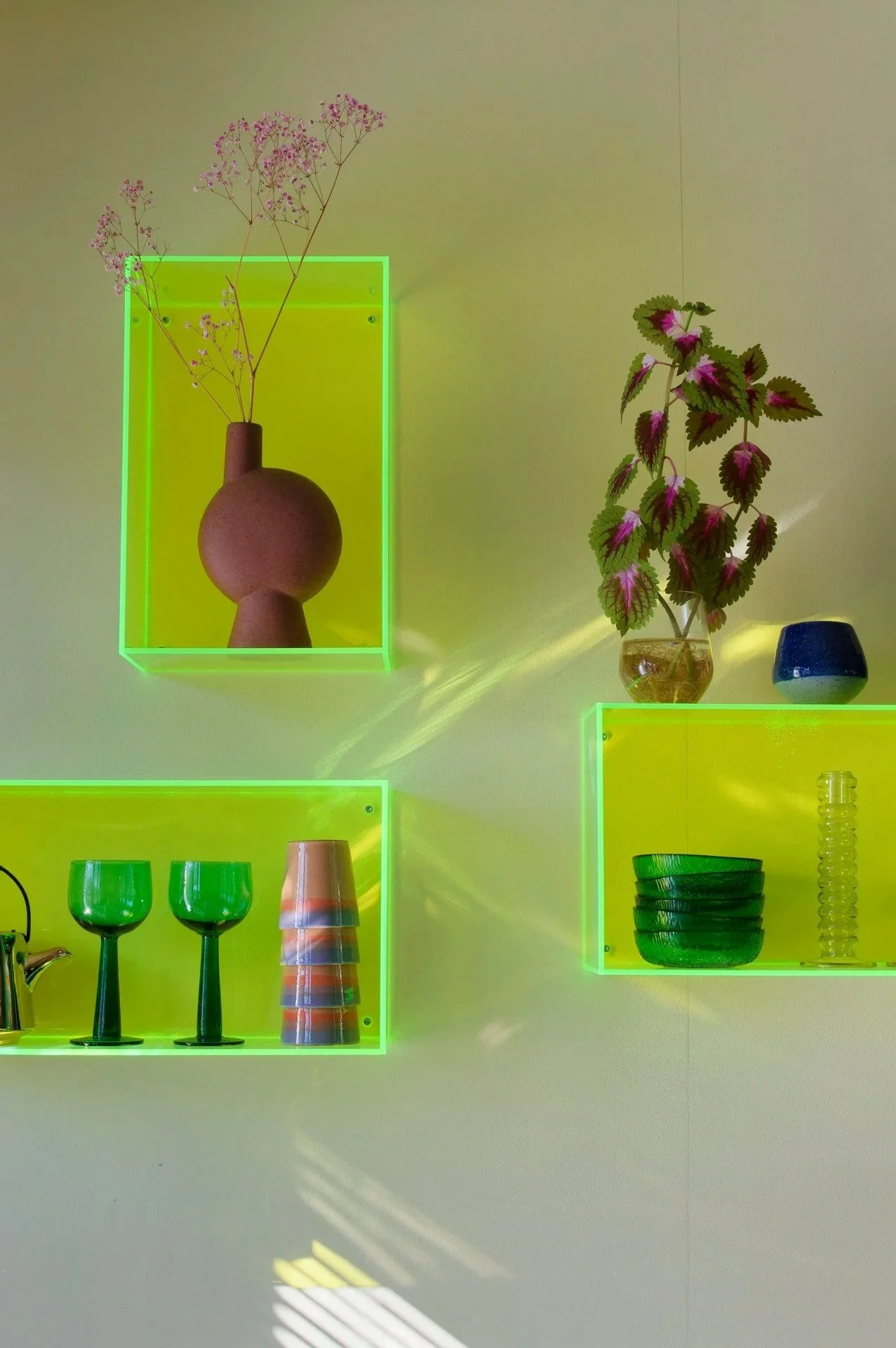 Decorative wall display with neon green framed shelves holding vases, plants, glasses, and bowls, illuminated by sunlight.