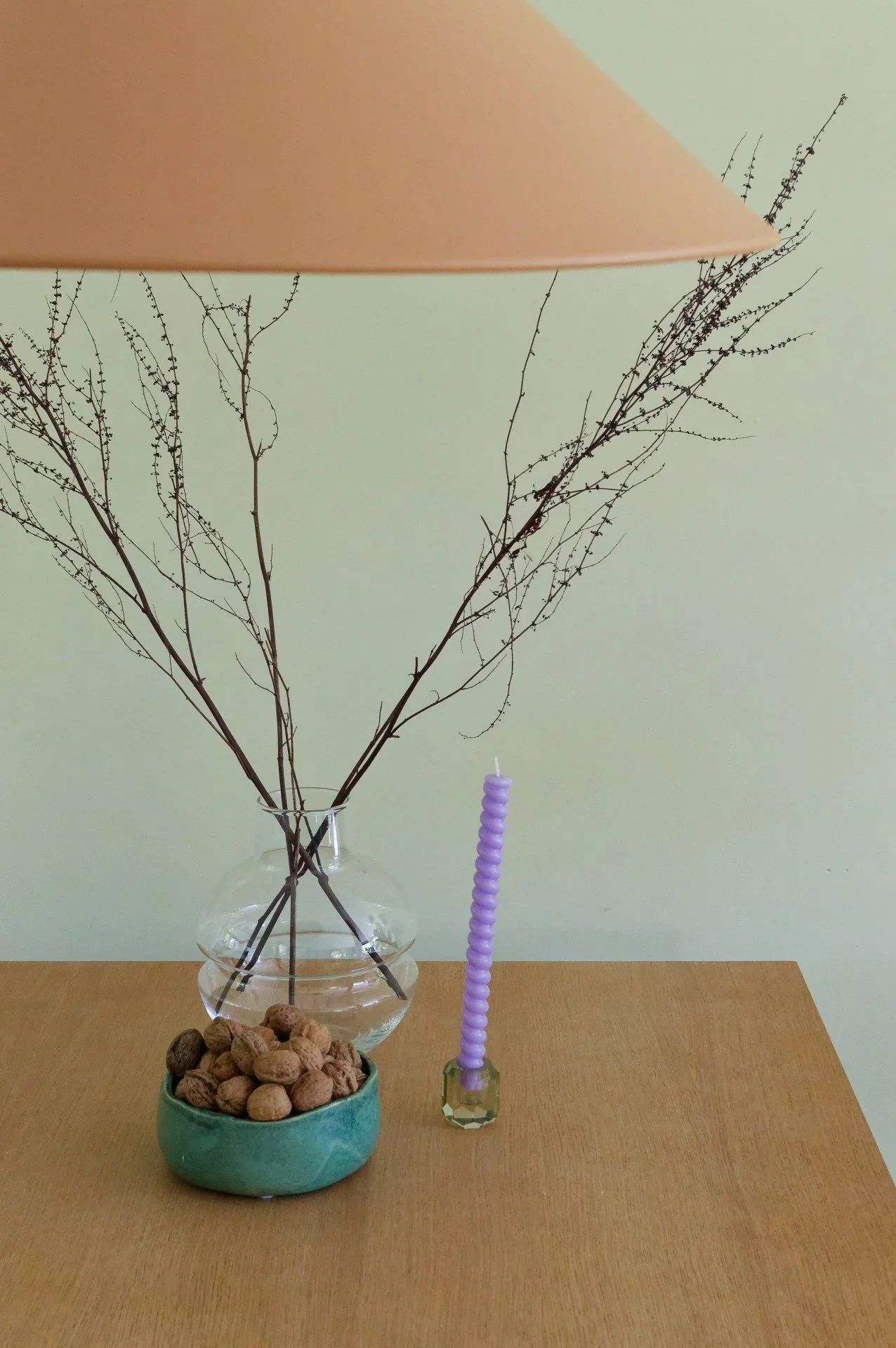 A wooden table with a glass vase holding dry twigs, a green bowl filled with walnuts, and a purple candle in a faceted glass holder, with a beige lampshade partially visible at the top.