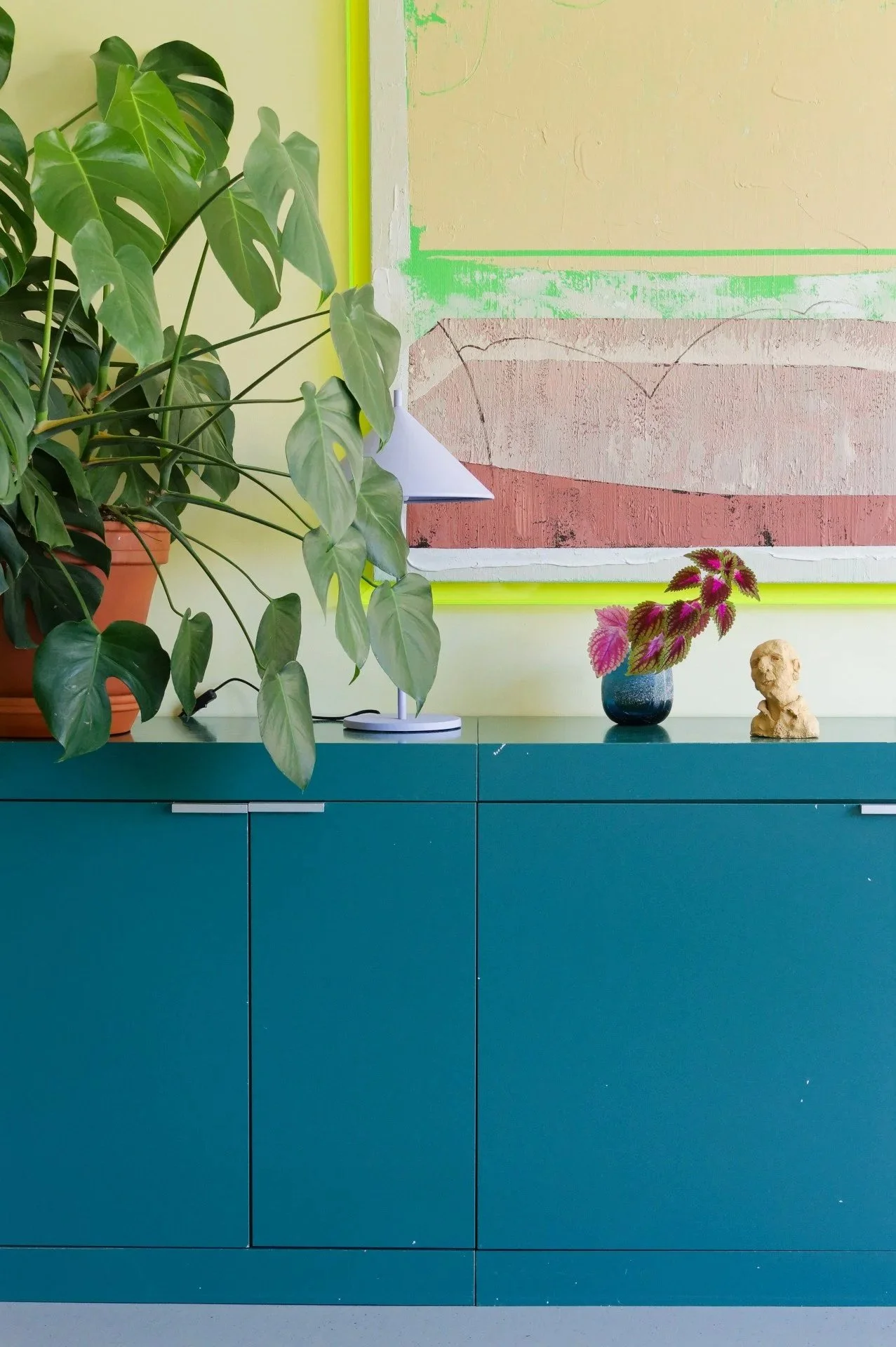 A teal-colored cabinet with a potted monstera plant, a modern white table lamp, a small black vase with pink and green leaves, and a small beige sculpture of a head, with a large paper artwork with pink, beige, and green colors hanging on a light green wall in the background.