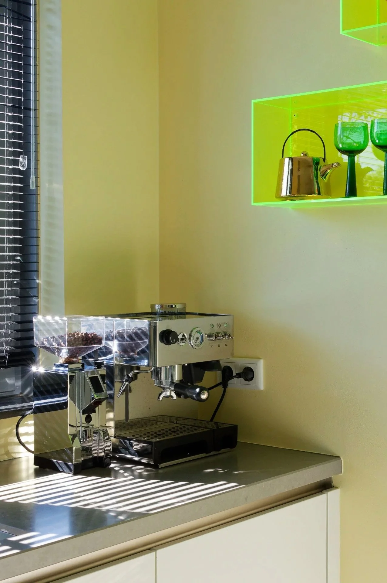 A modern coffee station with a metallic espresso machine on a beige countertop, illuminated by natural light through window blinds. Two glass containers filled with coffee beans are on the espresso machine, and there are neon green shelves on the wall holding a gold kettle and green glassware.