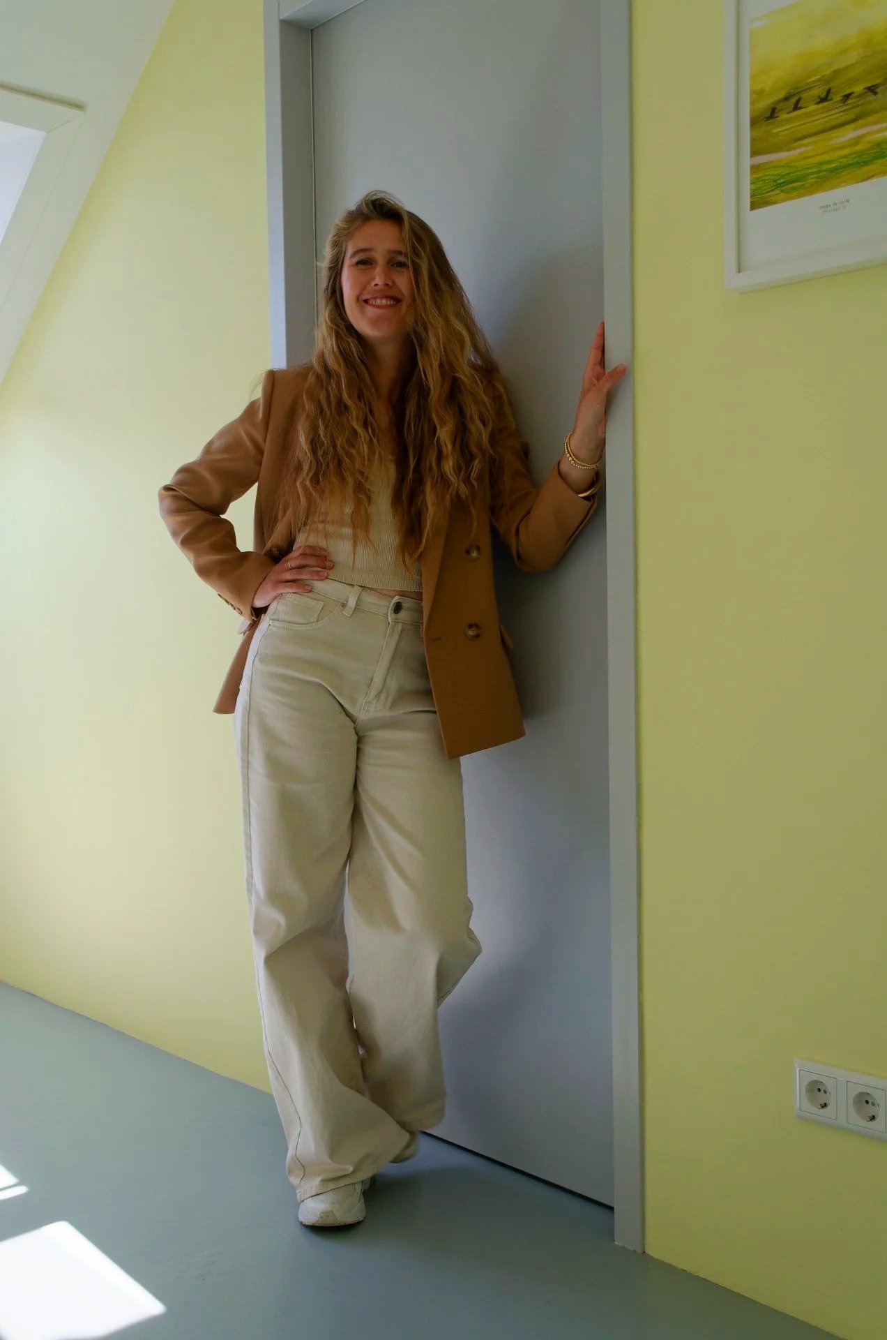 A woman with long blonde hair, dressed in beige pants, a beige sweater, and a tan blazer, stands in front of a closed door in a room with yellow and white walls, smiling at the camera.